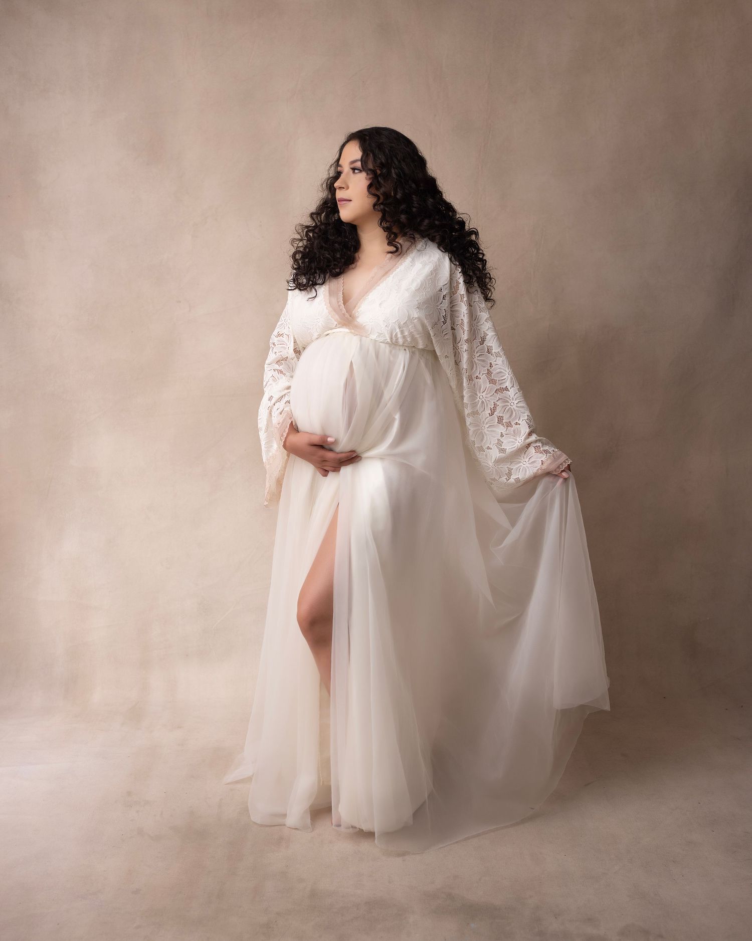Ethereal maternity portrait in flowing white lace gown against neutral backdrop.