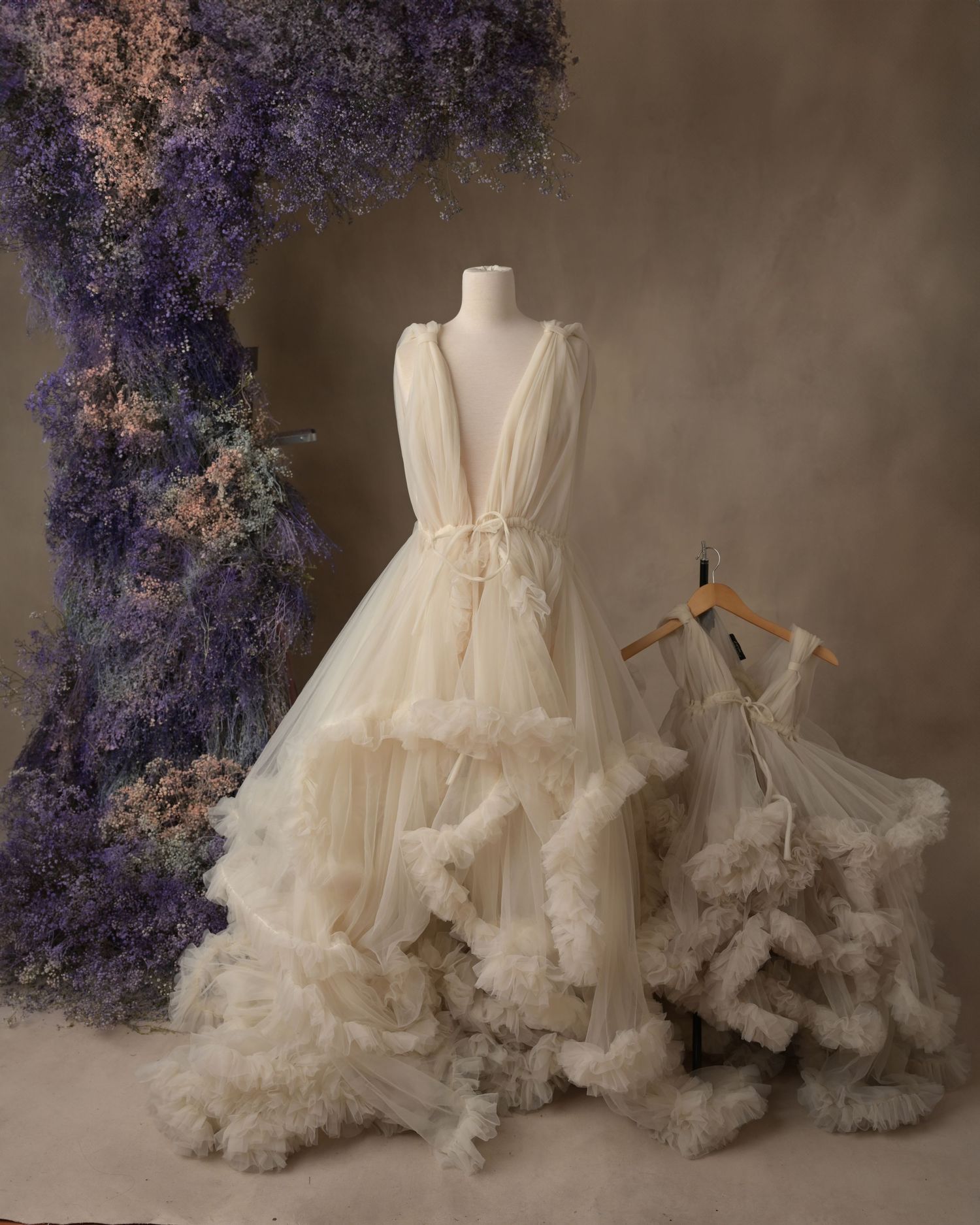 Elegant white tulle wedding gown with ruffled layers and a companion smaller dress displayed against purple floral decor.