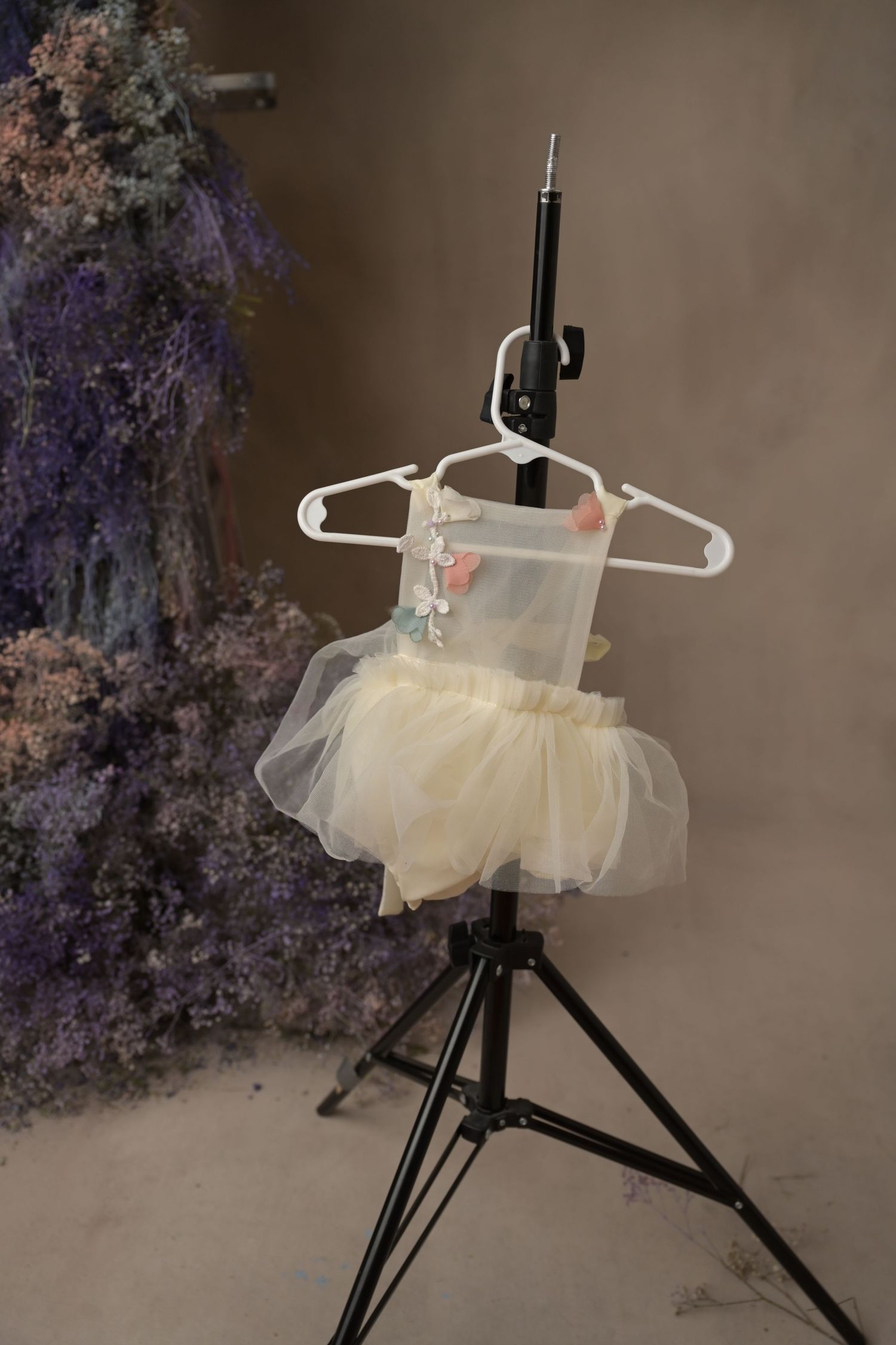 Small cream tulle dress displayed on a black lighting stand with delicate ruffles and floral backdrop.