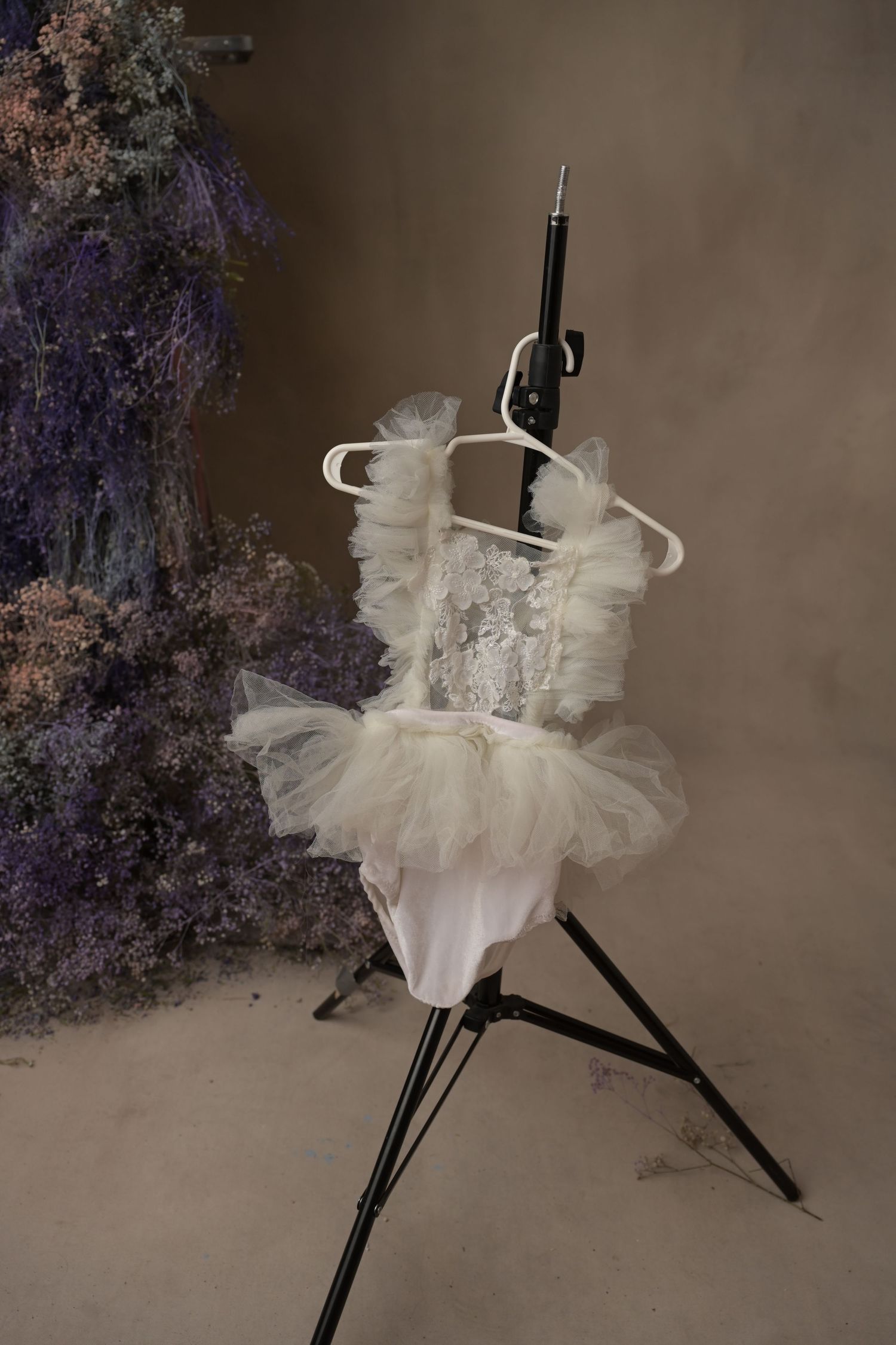 White tutu-style dress hanging on a photography light stand against a dark background with purple flowers.
