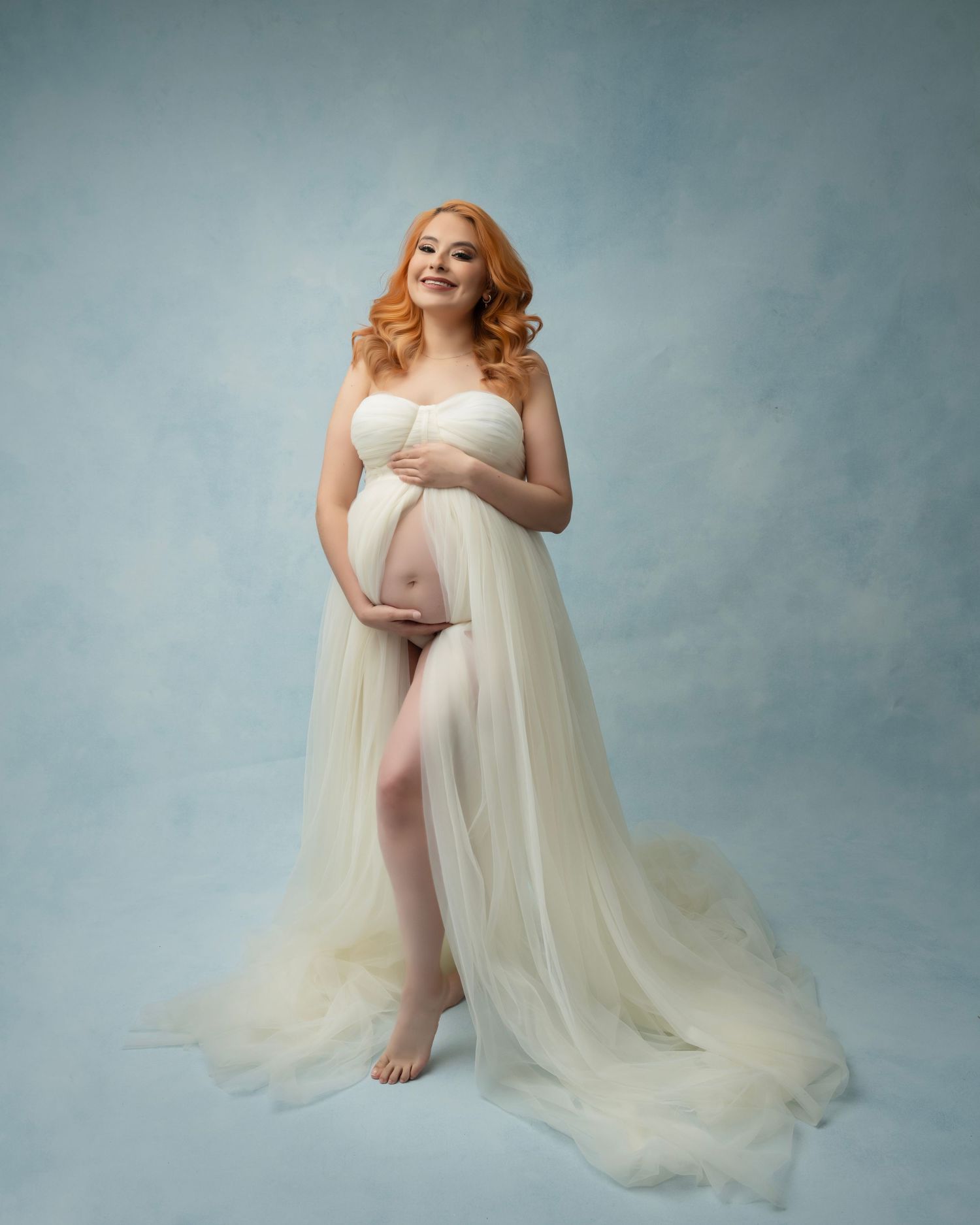 Elegant maternity photo featuring flowy cream gown against blue backdrop.