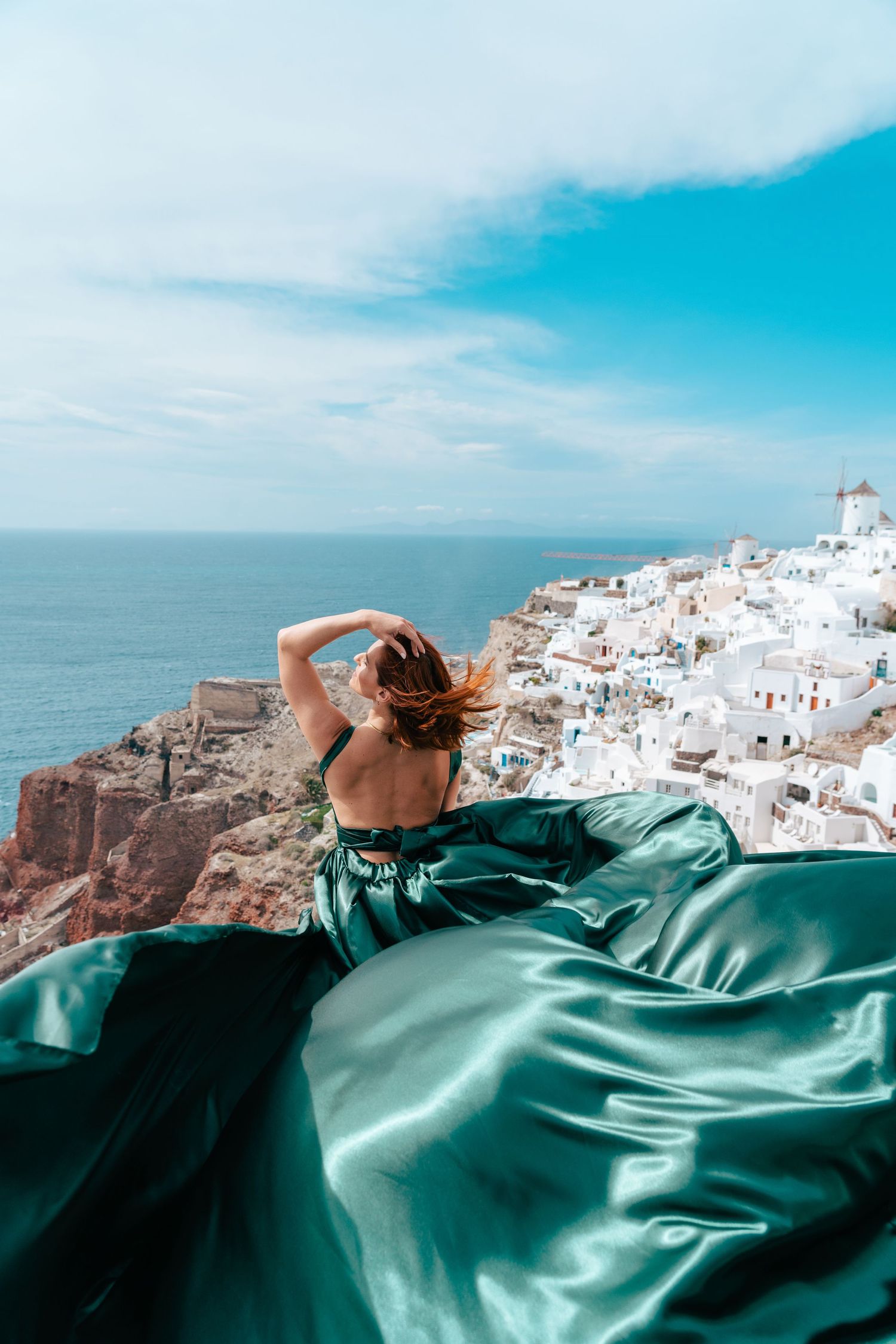 Flowing emerald green dress billows in the wind against the white buildings and blue waters of Santorini, Greece.
