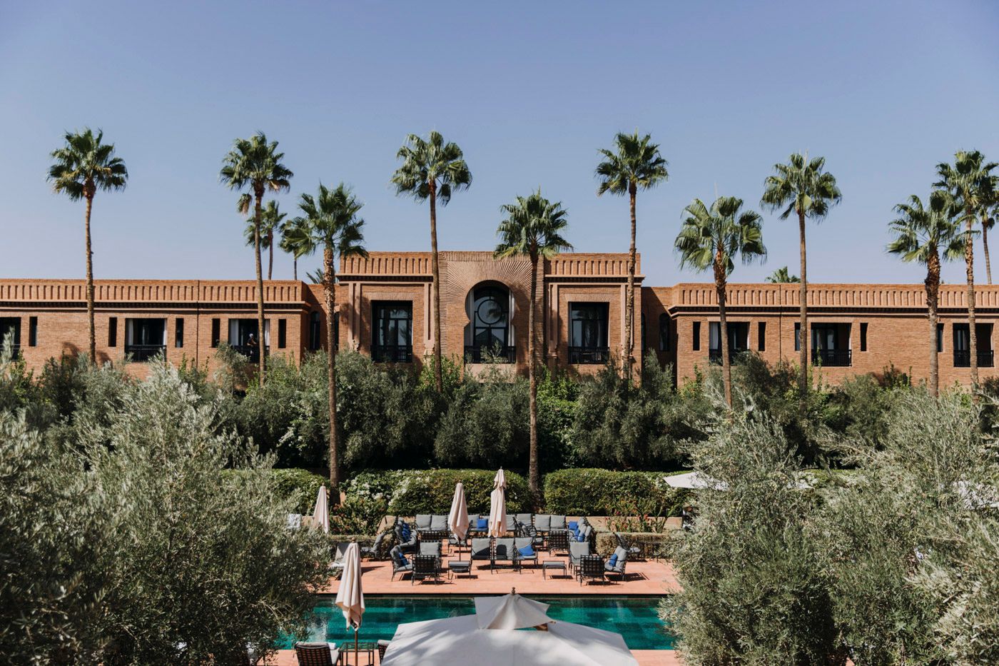 View of Mediterranean style resort with terracotta buildings, palm trees and pool surrounded by olive trees.