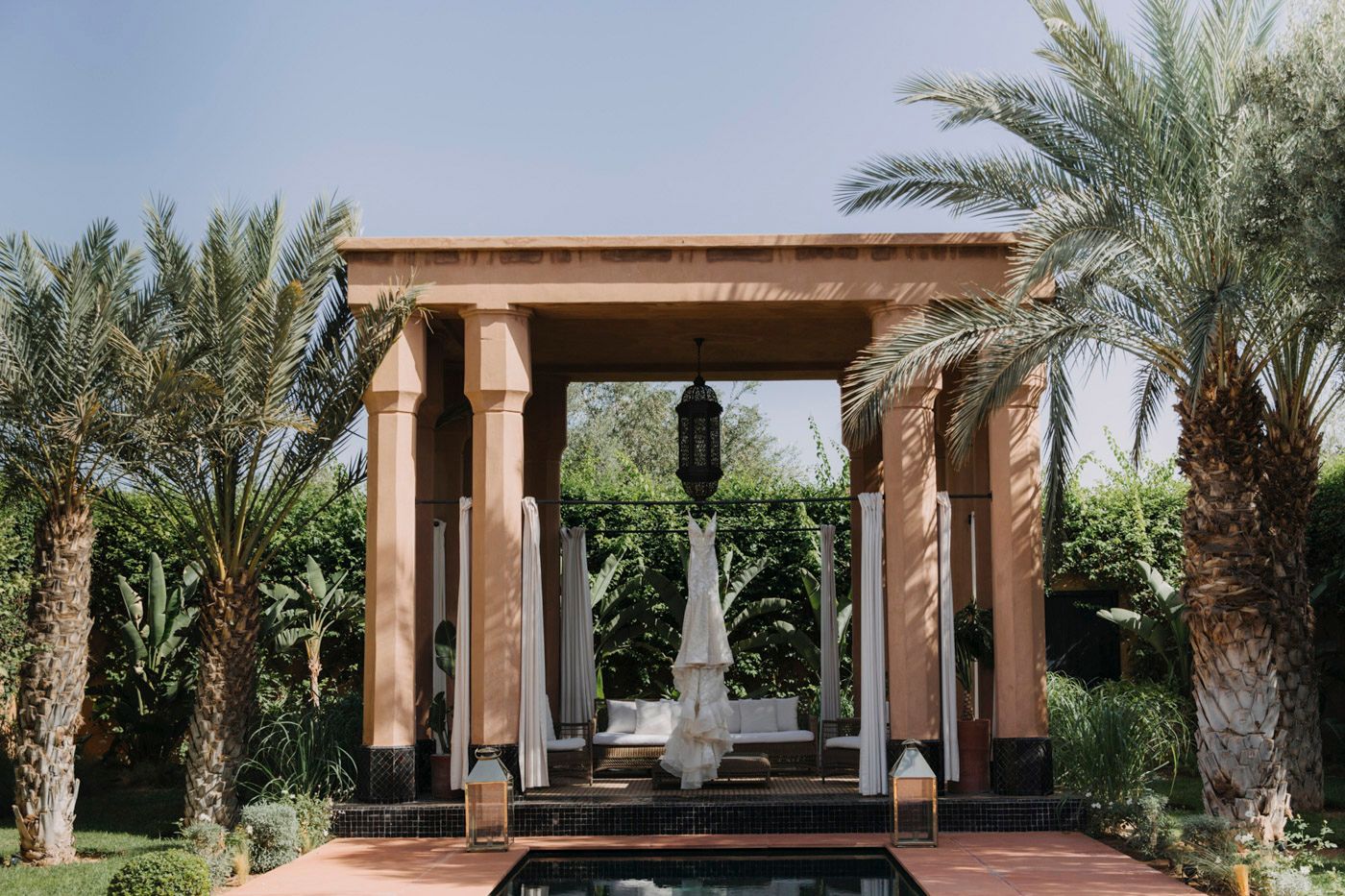 Mediterranean-style pergola with stone columns surrounded by palm trees and a reflecting pool in a garden setting.