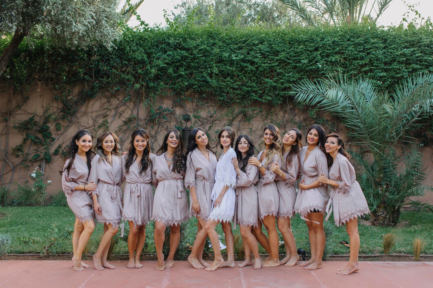 A group of bridesmaids wearing light pink dresses pose together in front of a lush garden wall.