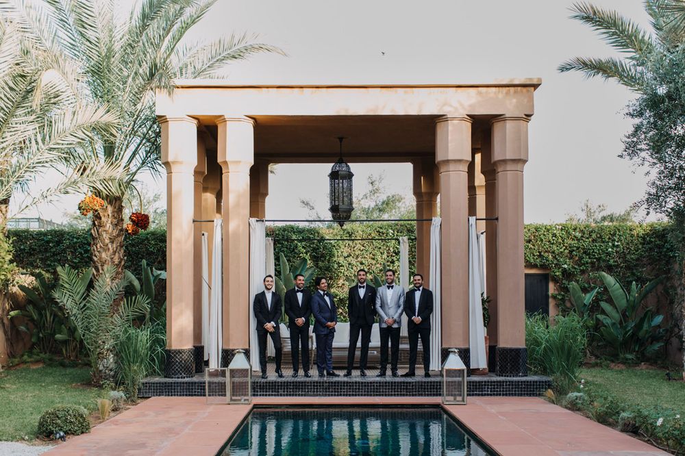 Formal wedding party poses under a columned gazebo next to a reflecting pool with palm trees.