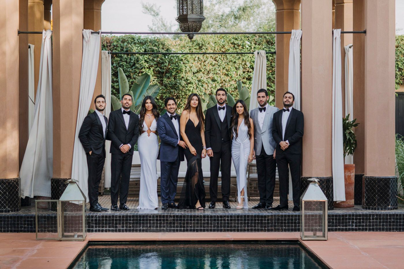 A formal group stands together in black and white attire on a patio platform beside a pool.