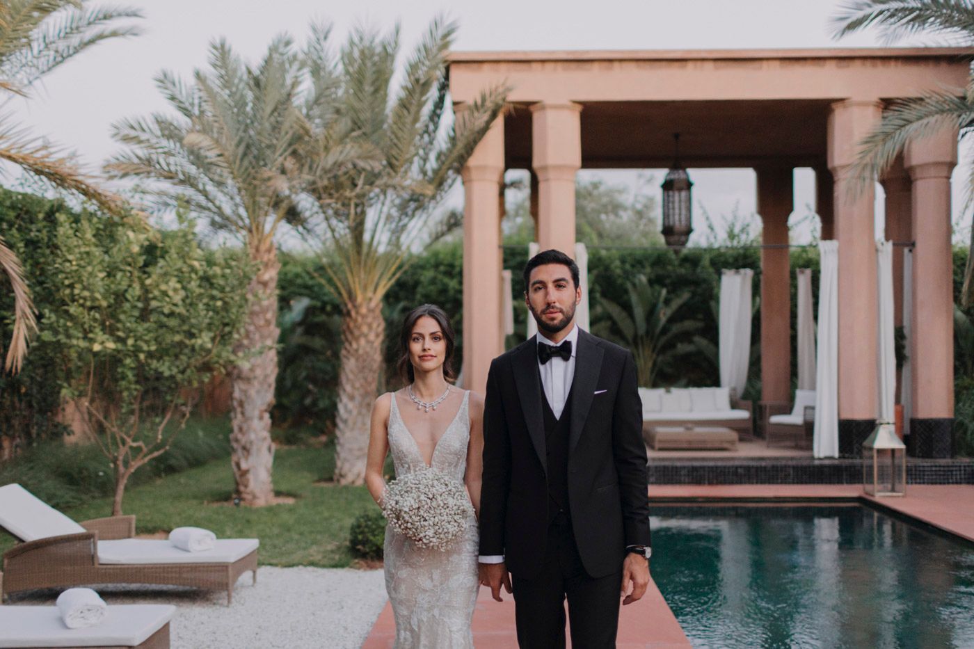 A couple in formal attire poses in front of a luxury villa pool with palm trees and pink-hued architecture.