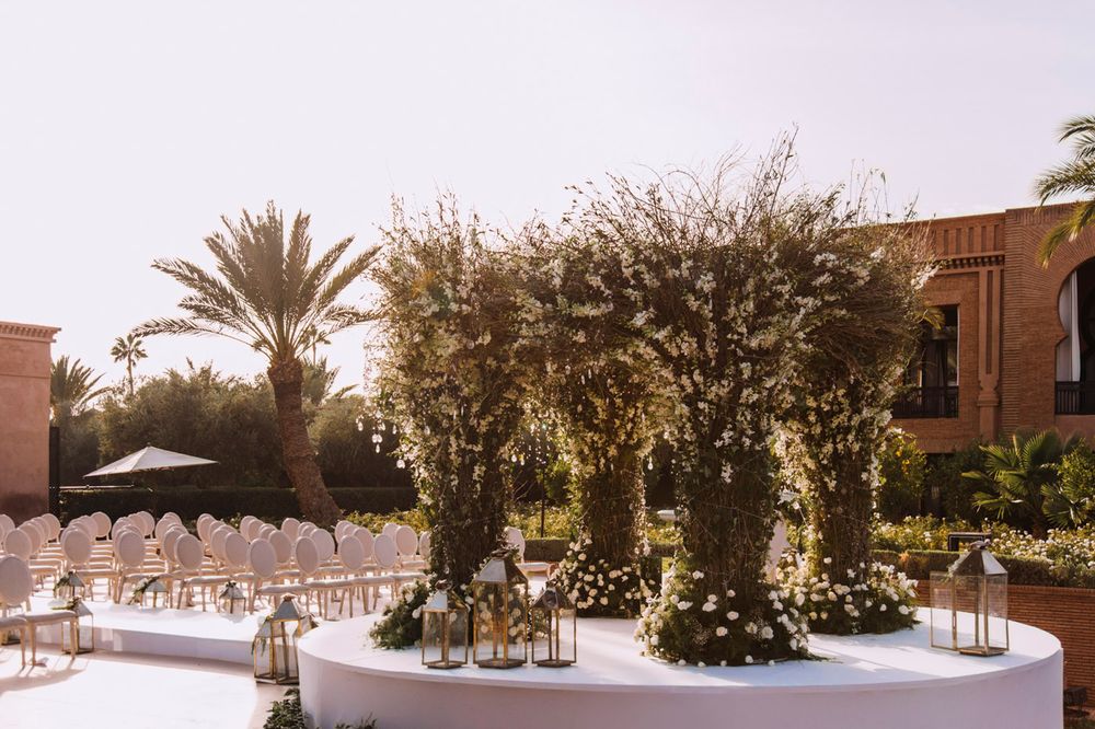 Elegant wedding reception setup with white tables and floral arrangements surrounded by palm trees at sunset.