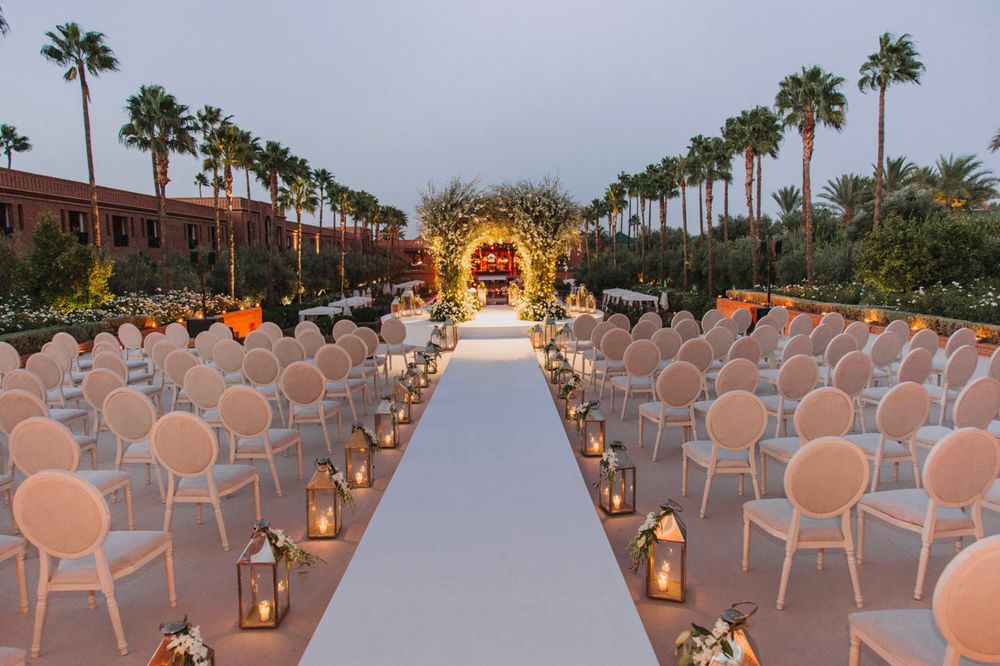 White chairs line a wedding aisle with lanterns leading to an illuminated floral arch under palm trees at dusk.
