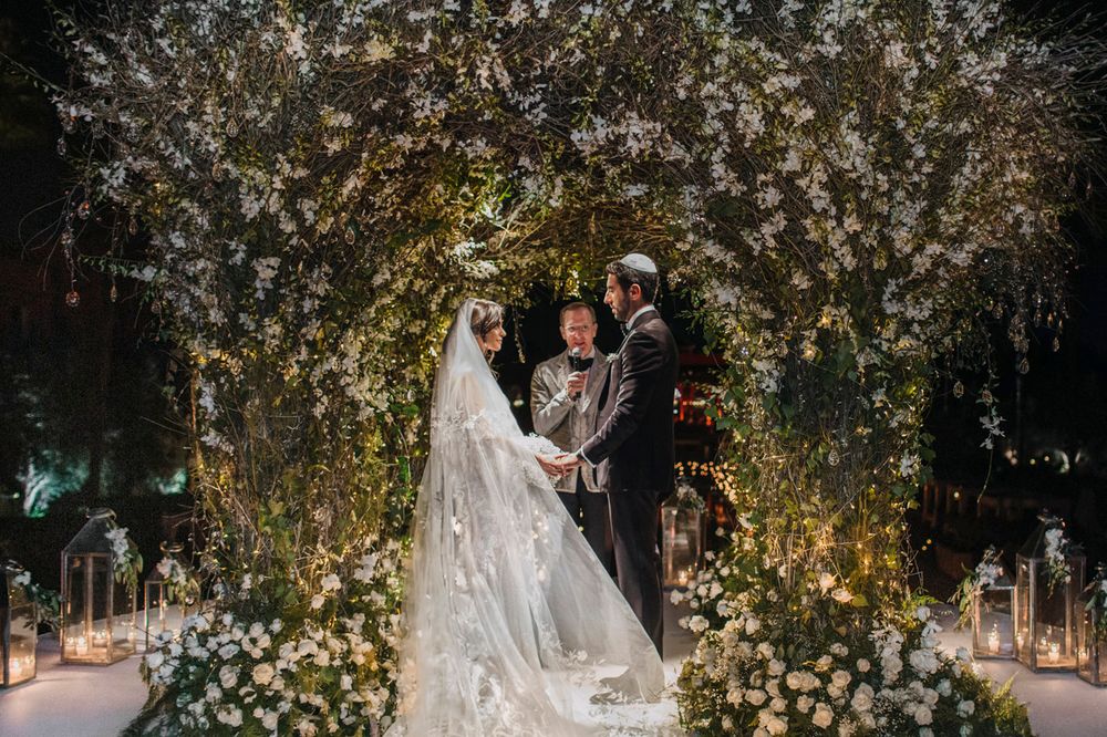 A wedding ceremony takes place under a magical archway of twinkling lights and white blossoms.