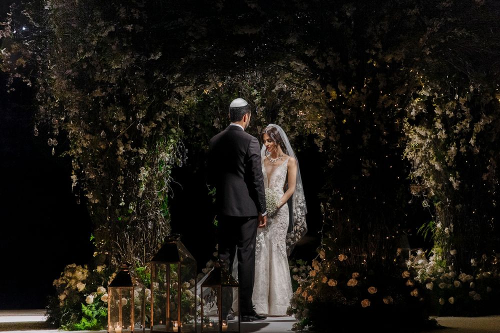 A couple stands beneath a romantic garden archway adorned with greenery and twinkling lights at dusk.