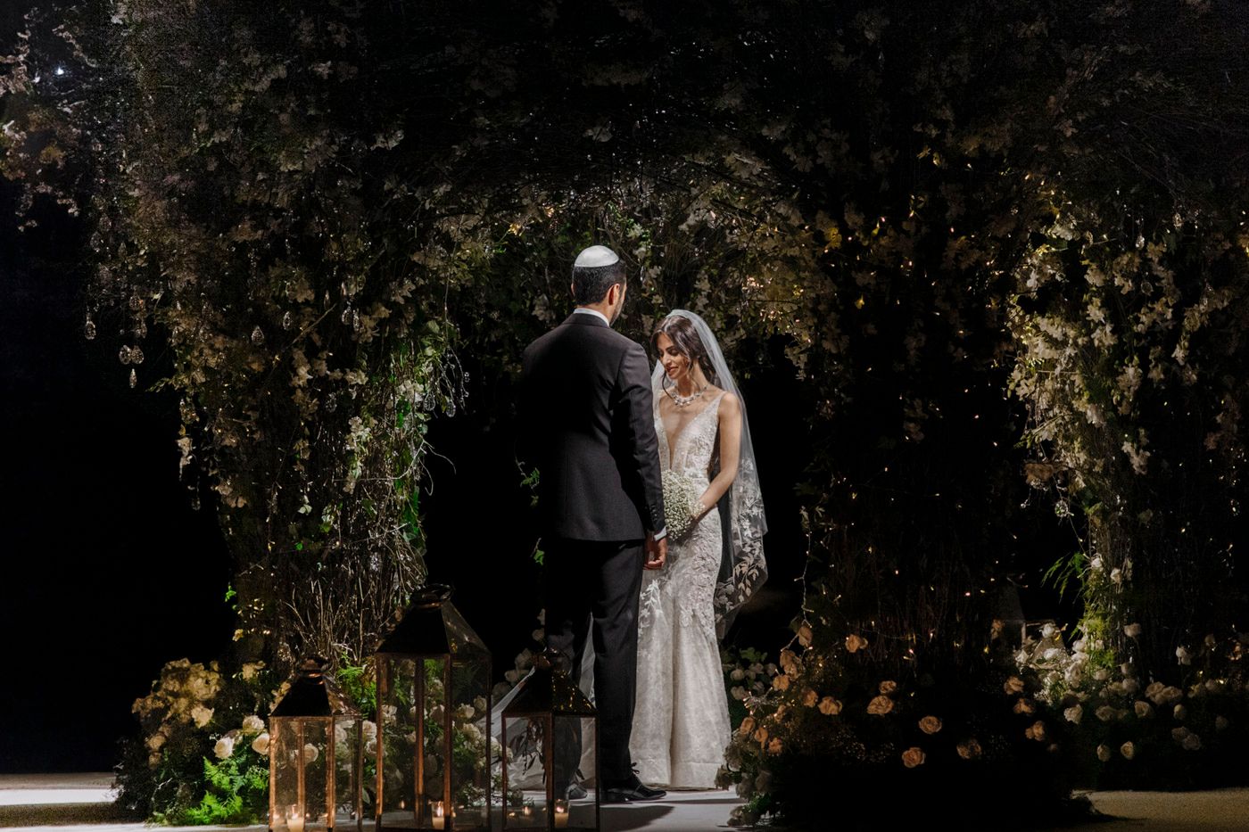A couple stands beneath a romantic garden archway adorned with greenery and twinkling lights at dusk.