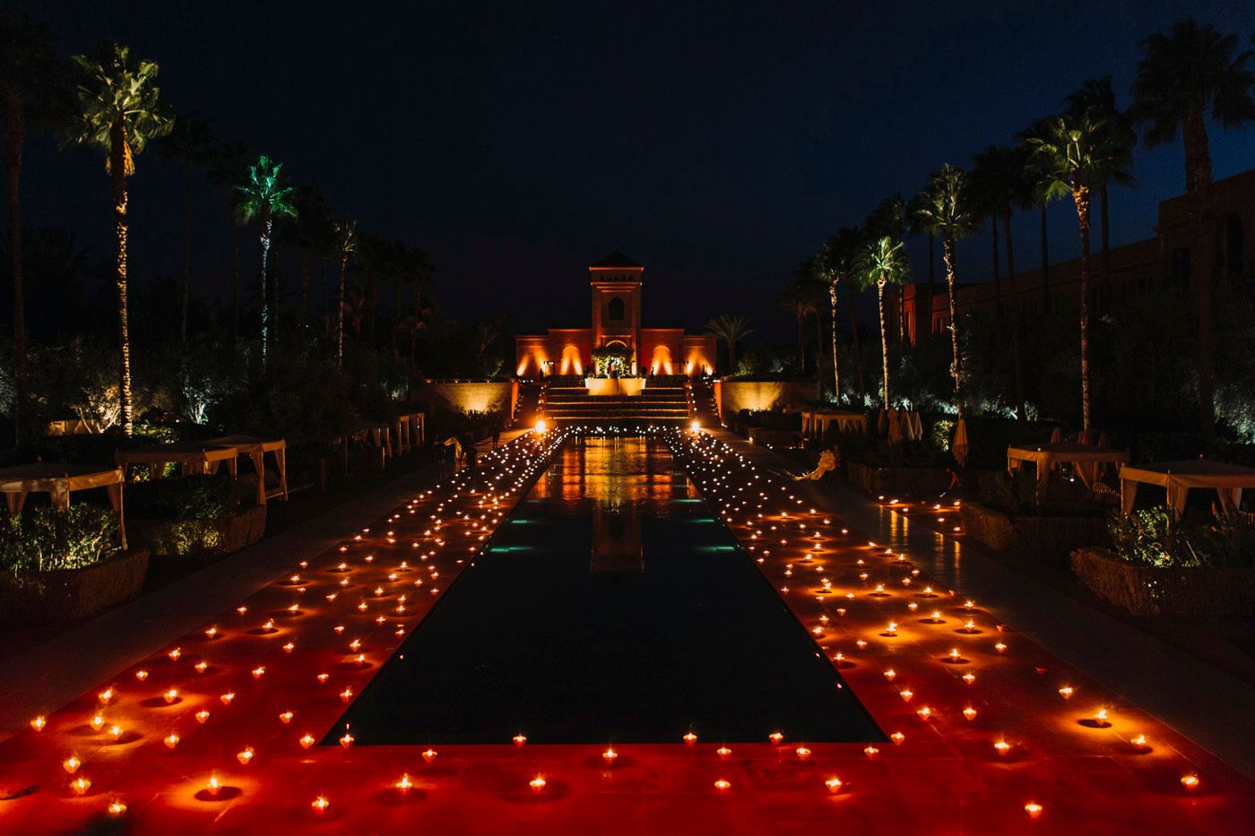 Palm trees and luminaries line a reflecting pool leading to an illuminated building at night during a special event.