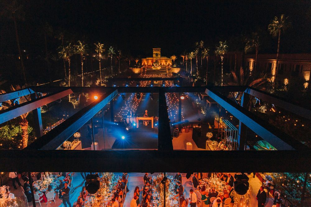 Illuminated outdoor wedding reception with glowing tables arranged around a pool under palm trees at night.