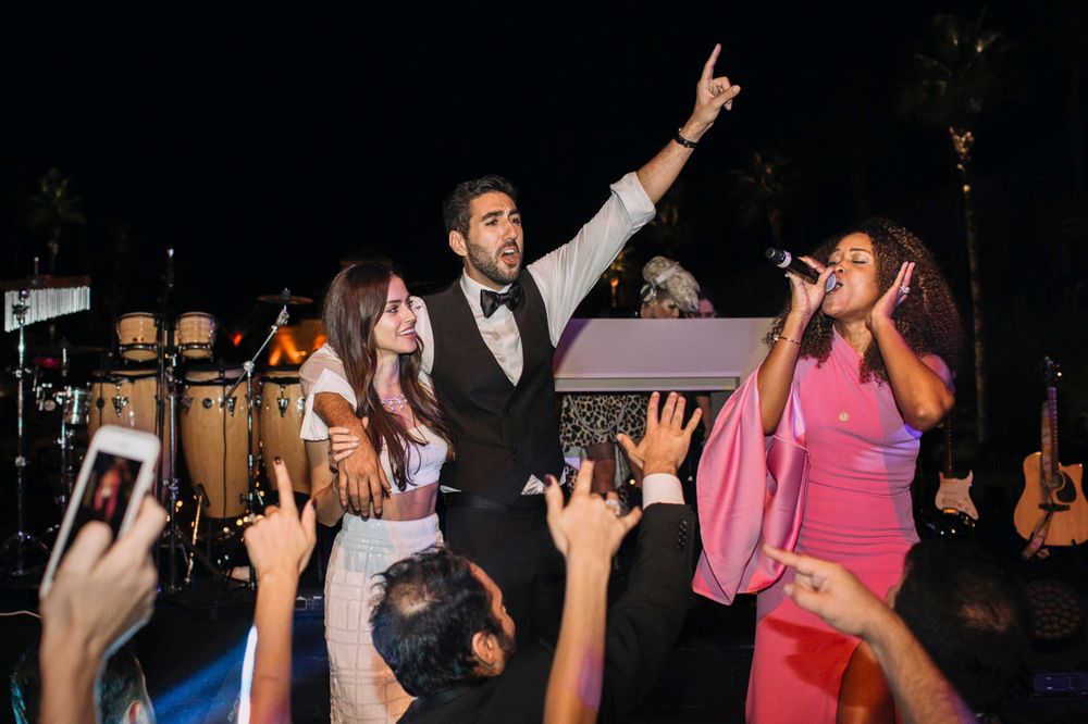 Energetic performers on stage with crowd raising hands at a lively nighttime event with instruments in the background.