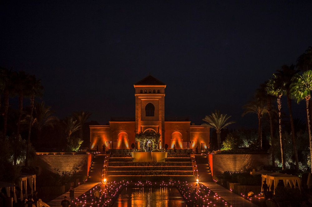 Egyptian-style building illuminated at night with orange lights reflecting in water and palm trees along pathway.