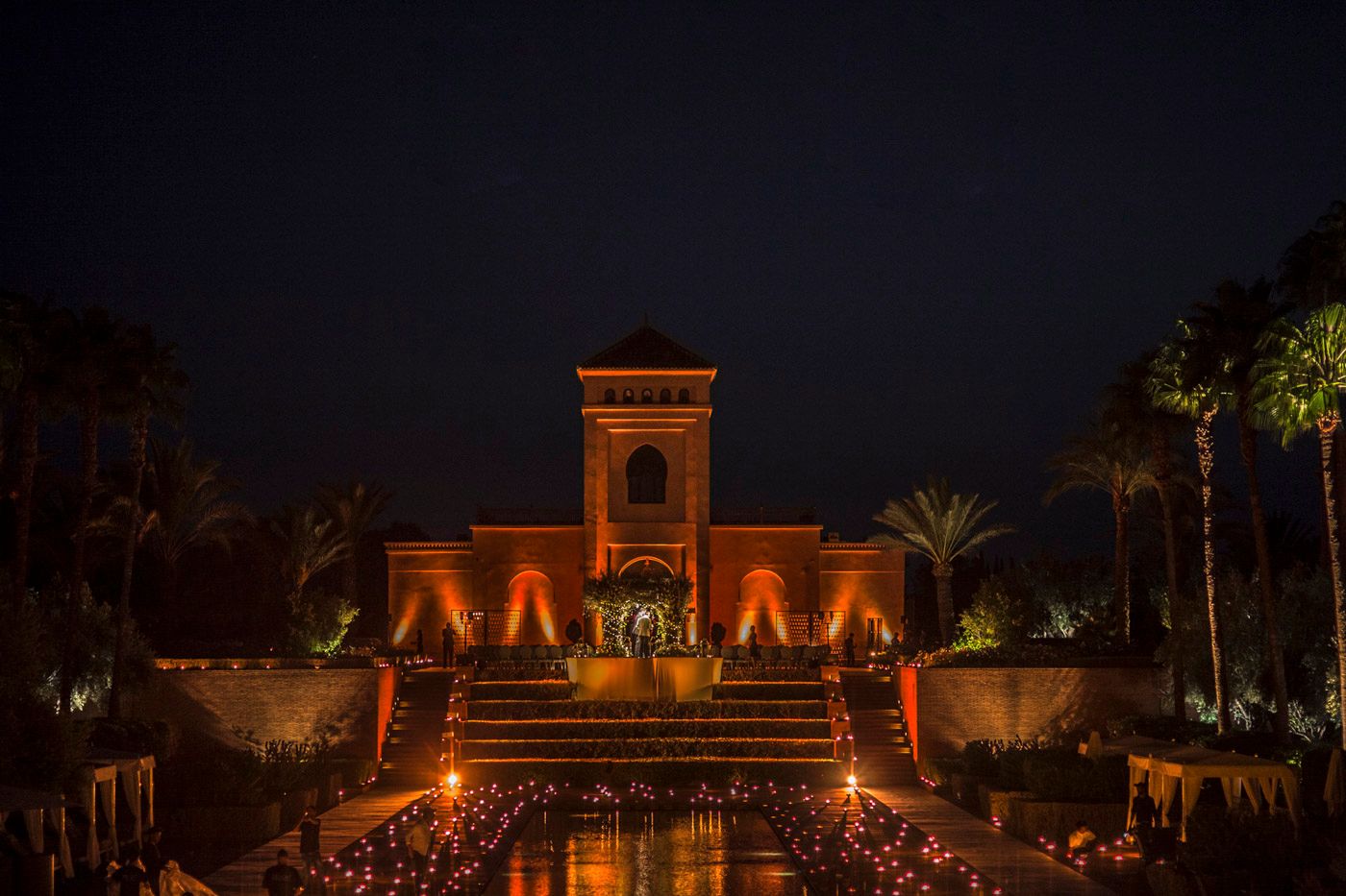 Egyptian-style building illuminated at night with orange lights reflecting in water and palm trees along pathway.