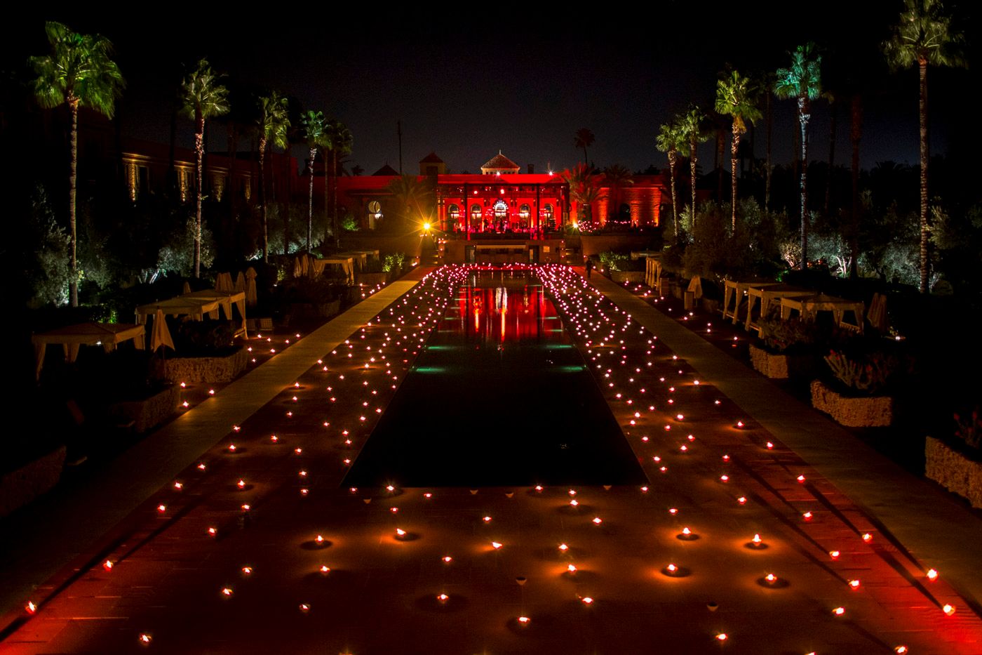A nighttime pathway lit by glowing luminaries leads to a red-illuminated building with palm trees lining both sides.