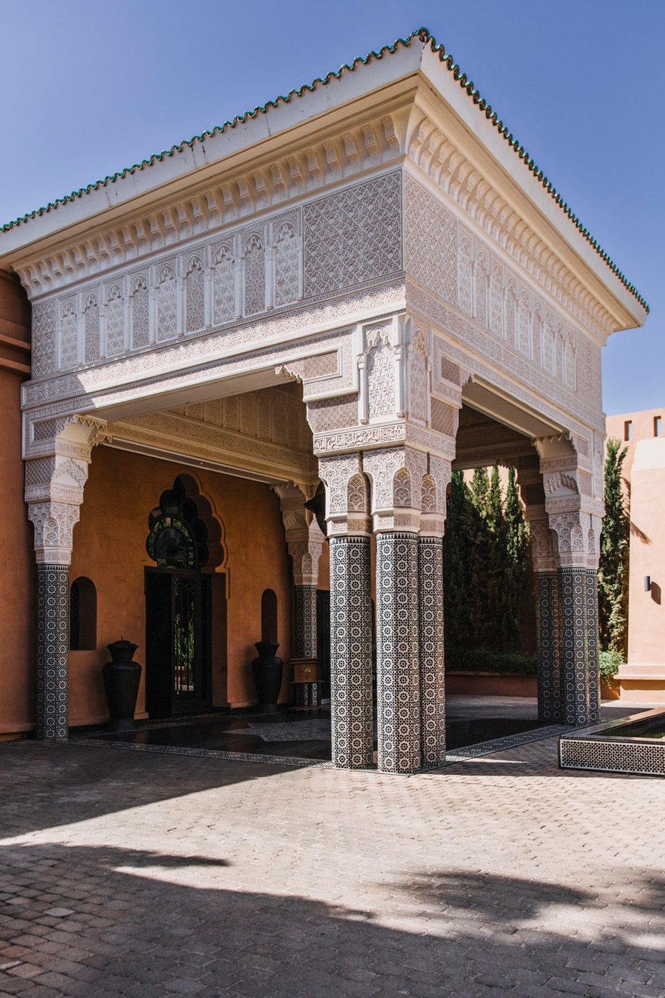 An ornate Moroccan-style entrance with white carved pillars, decorative tile work and arabesque patterns against a blue sky.