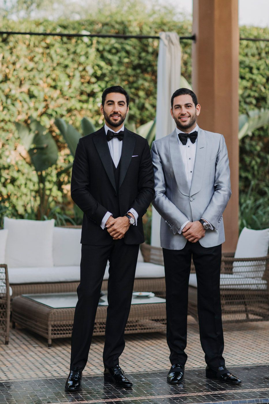 Two men stand in formal attire - one in black tuxedo and one in light gray suit jacket - at an outdoor venue.