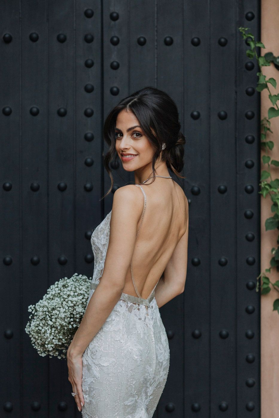 Elegant bride in form-fitting lace backless wedding gown holds baby's breath bouquet against dark rustic door.
