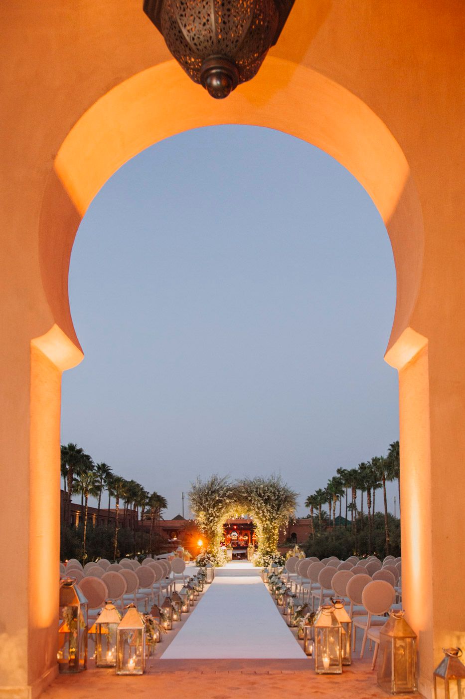 Elegant outdoor wedding ceremony setup with glowing lanterns and white chairs viewed through an orange archway at dusk.