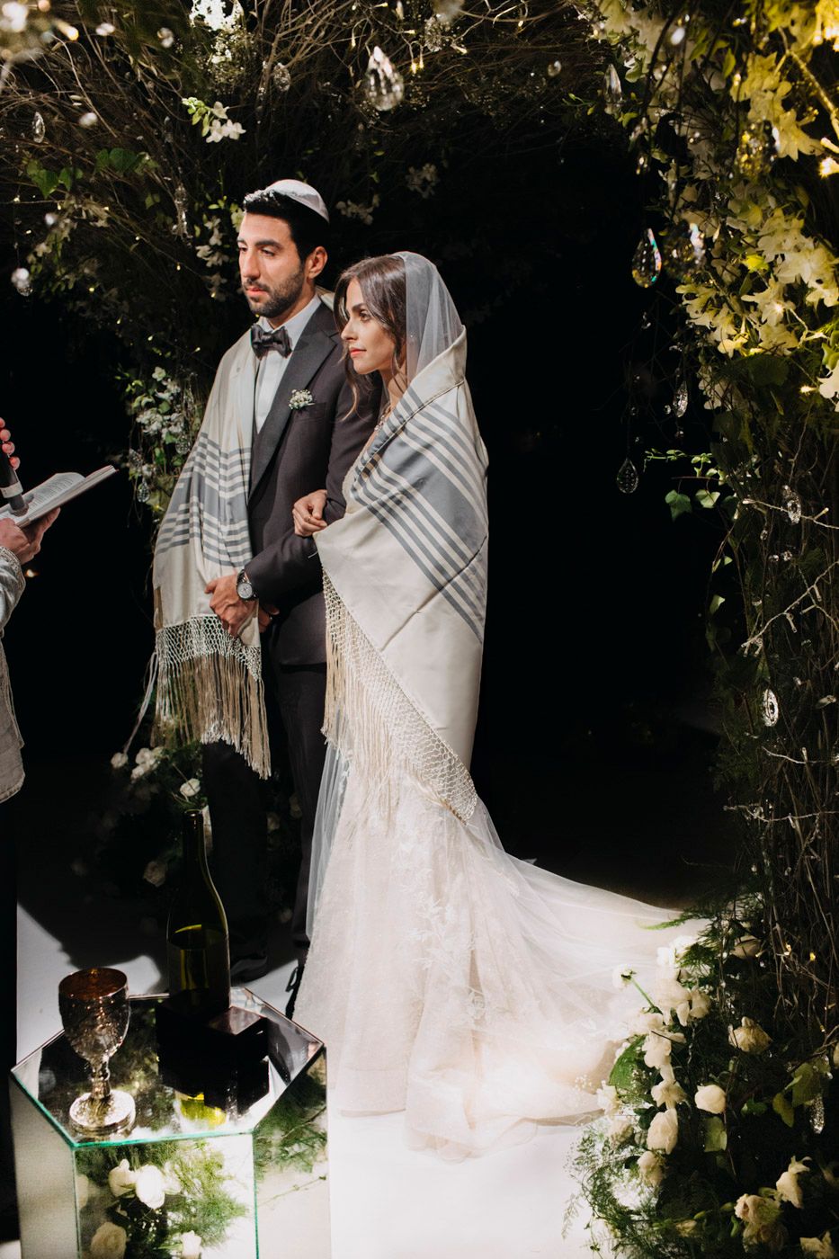 A couple stands under a floral archway during a Jewish wedding ceremony with traditional prayer shawls and wine glass.