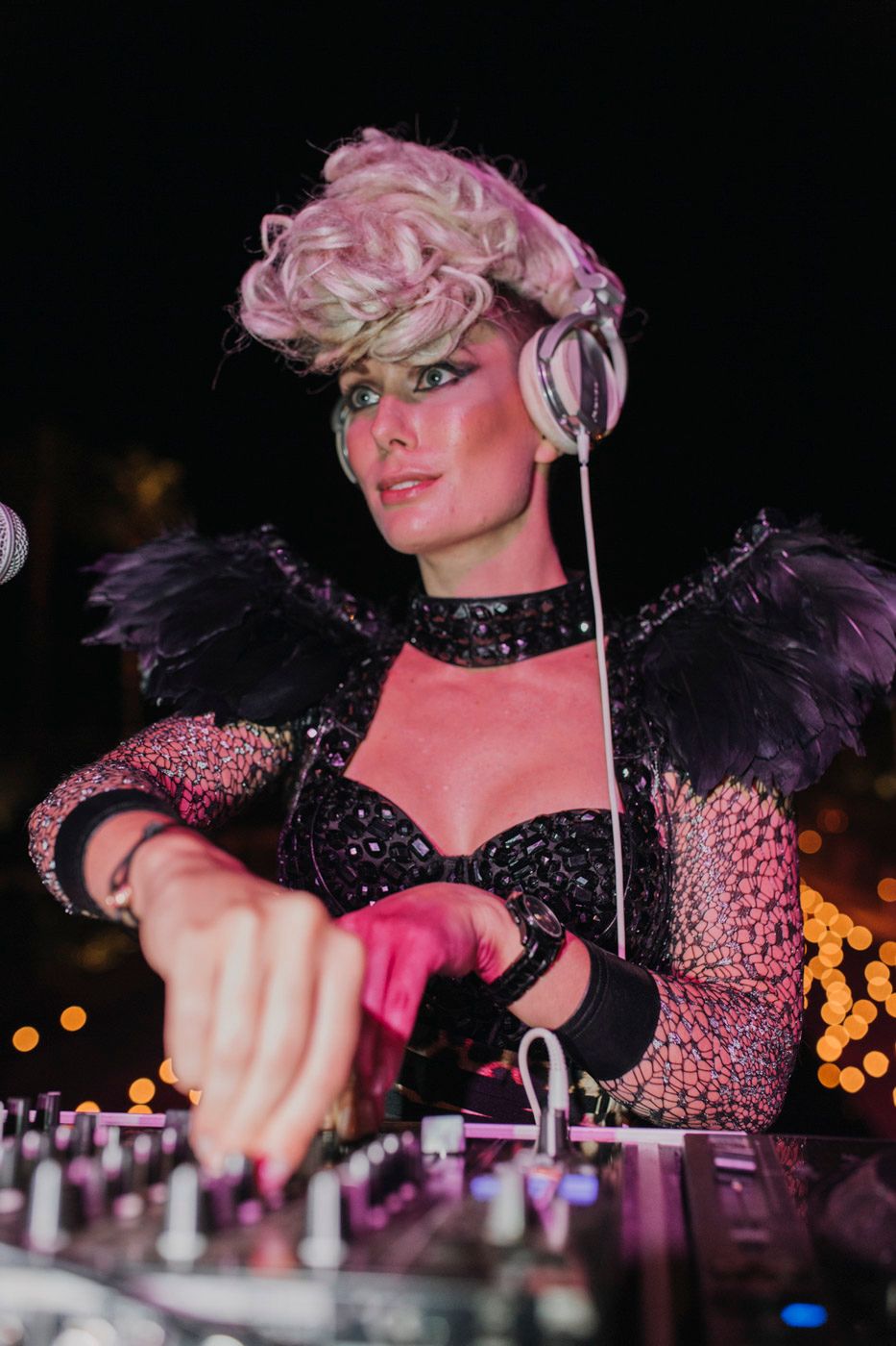 An entertainer in a glittering black costume with feathers and pink hair performs at a DJ booth during a nightclub event.