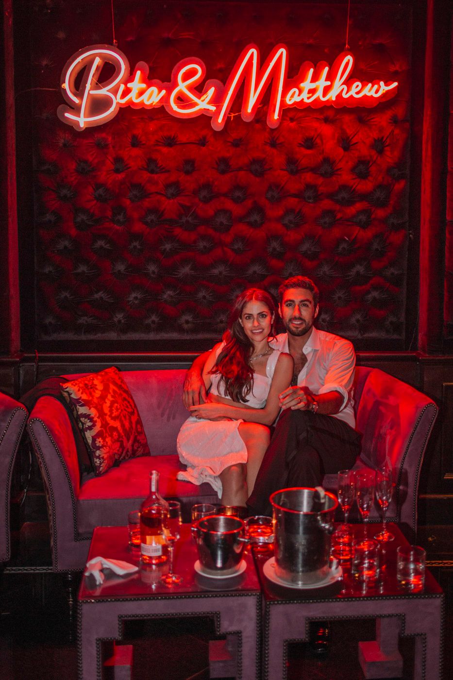 A couple sits on a red velvet couch beneath a glowing neon sign in a dimly lit lounge setting.