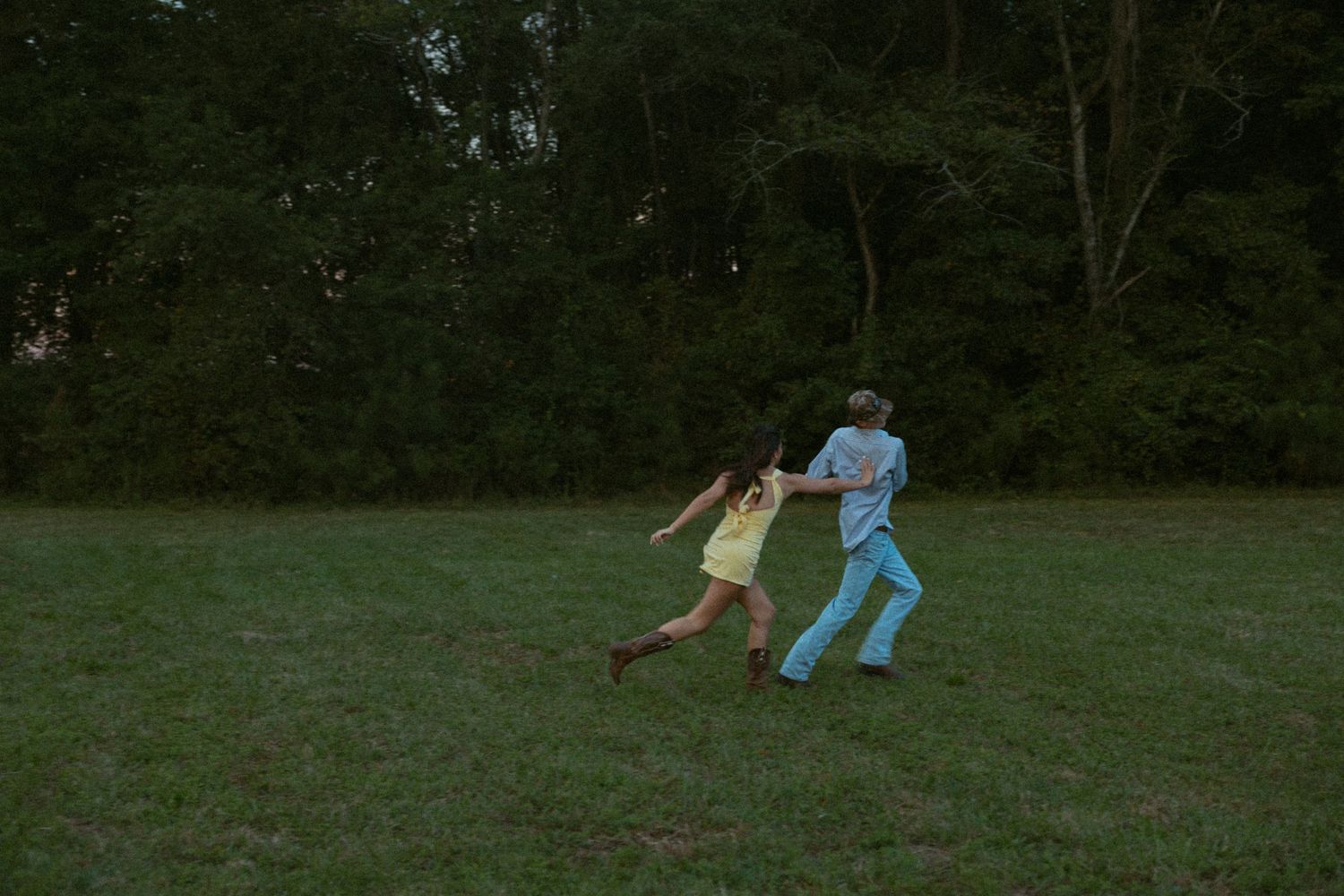Two people run playfully through a grassy field at dusk.