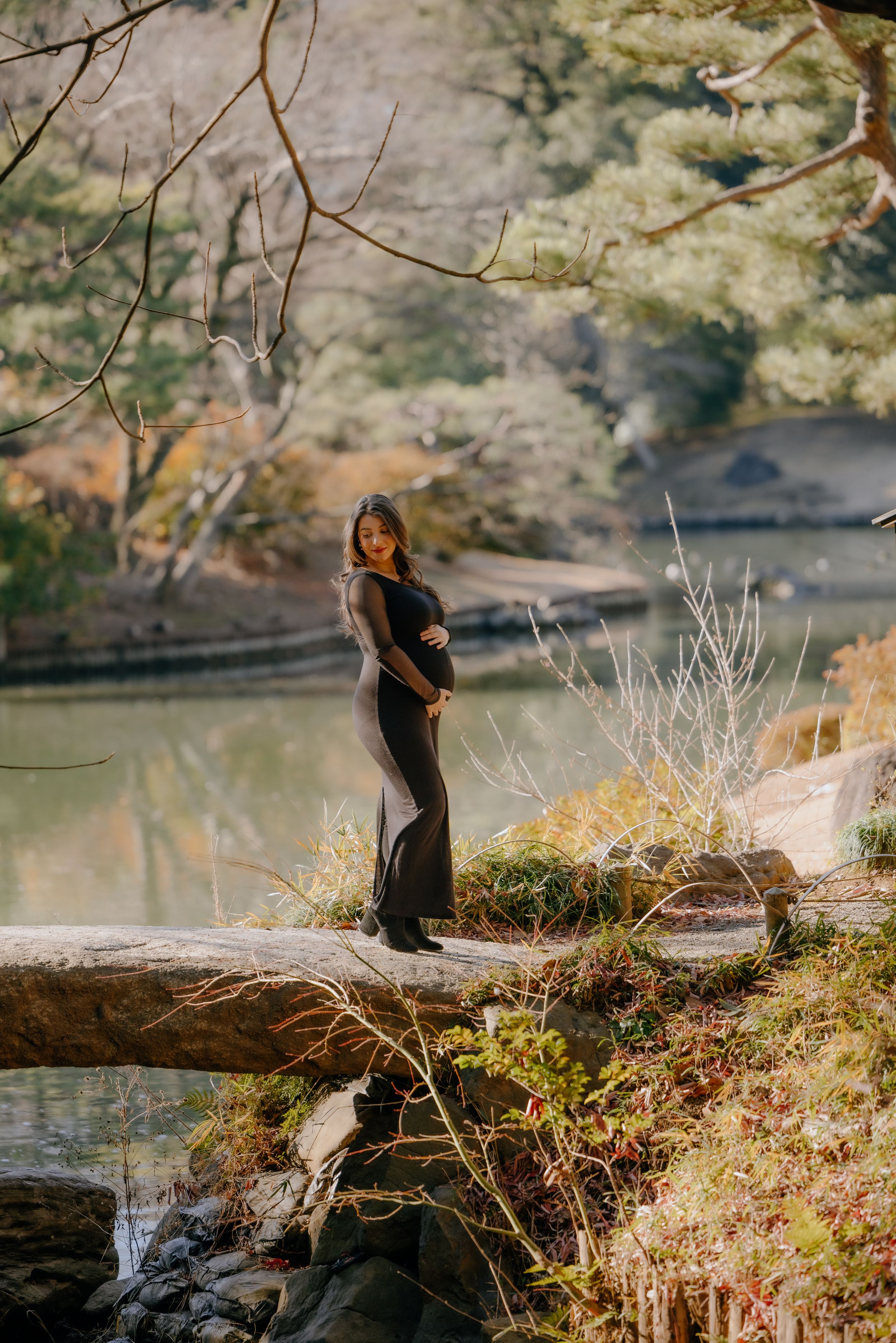 A pregnant woman in black stands on a fallen log by a tranquil pond during autumn.