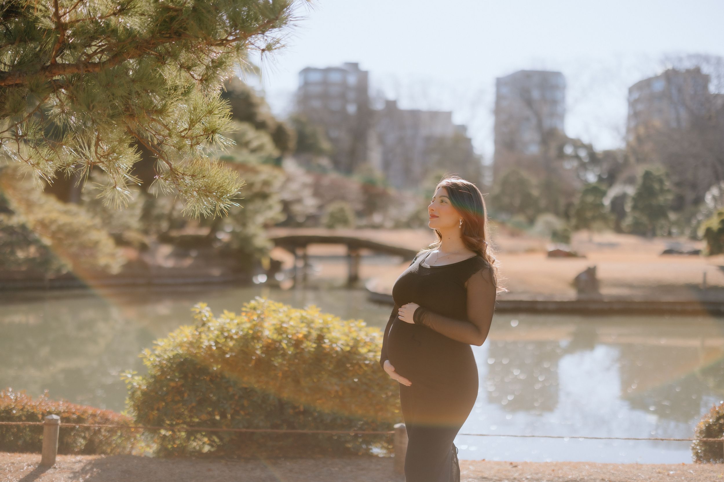 Pregnant woman in black dress stands beside lake in urban park during golden hour with cityscape in background.