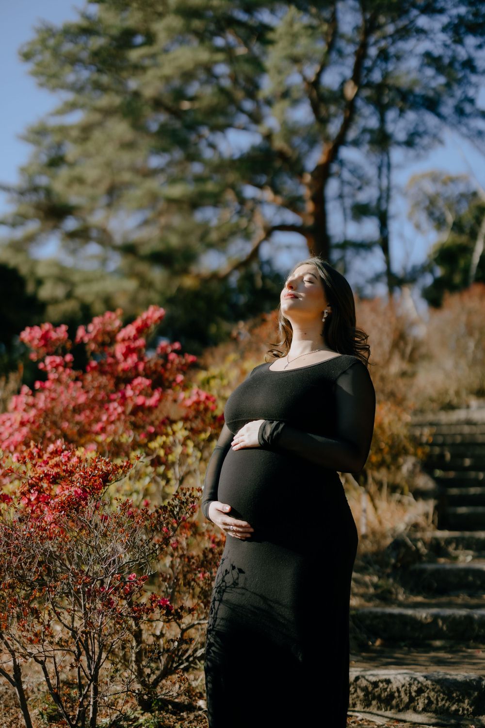 Elegant maternity silhouette in black dress poses in garden with pink flowers and sunlit trees in background.