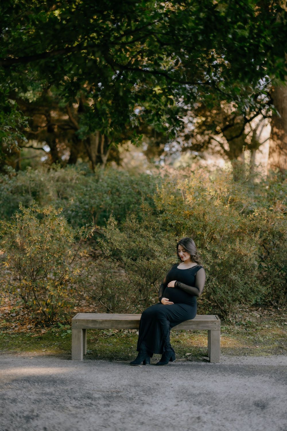 A pregnant woman in a dark dress sits contemplatively on a wooden bench along a path surrounded by greenery at sunset.
