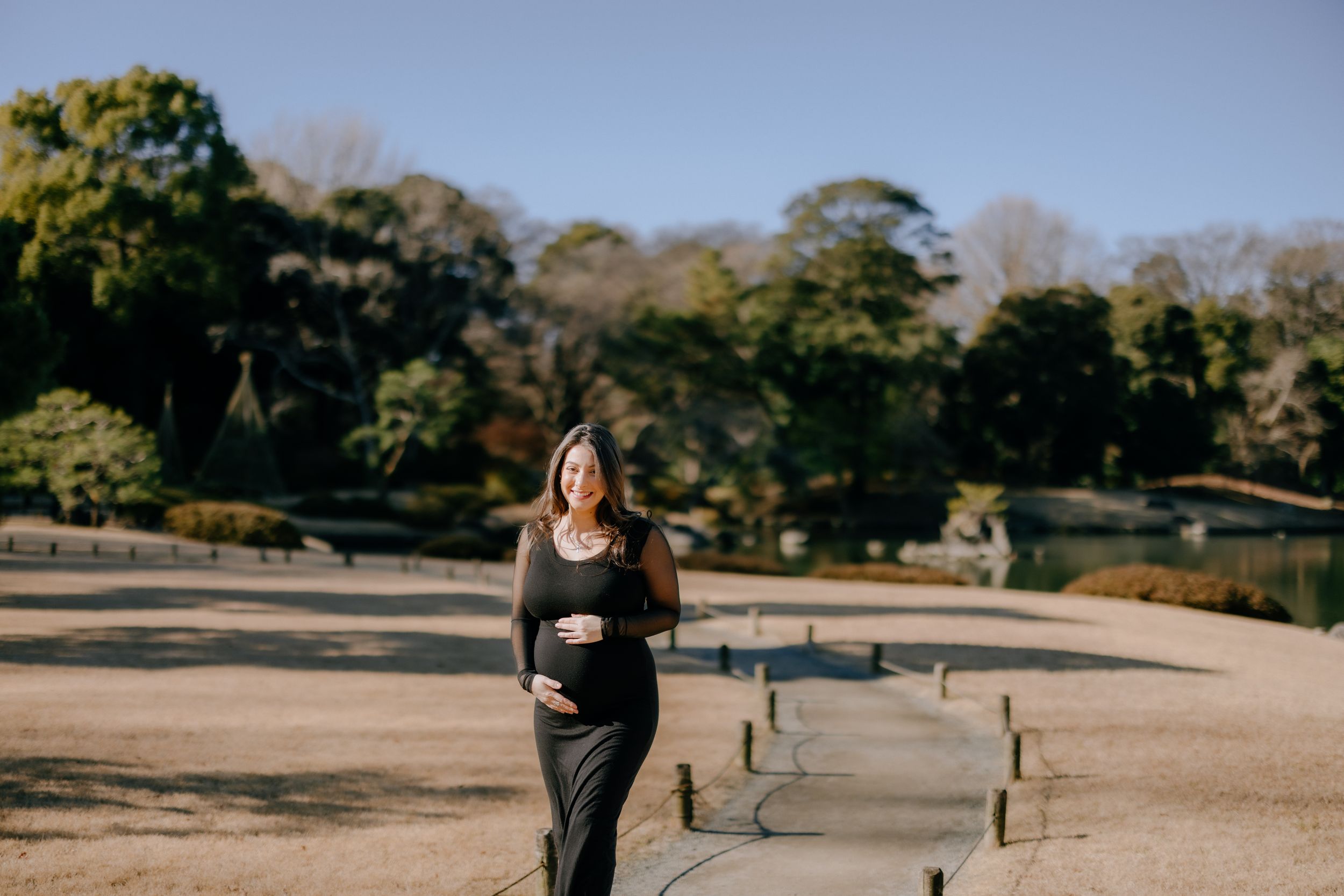 A person wearing a black dress stands on a dirt path in a park with trees in the background on a sunny day.