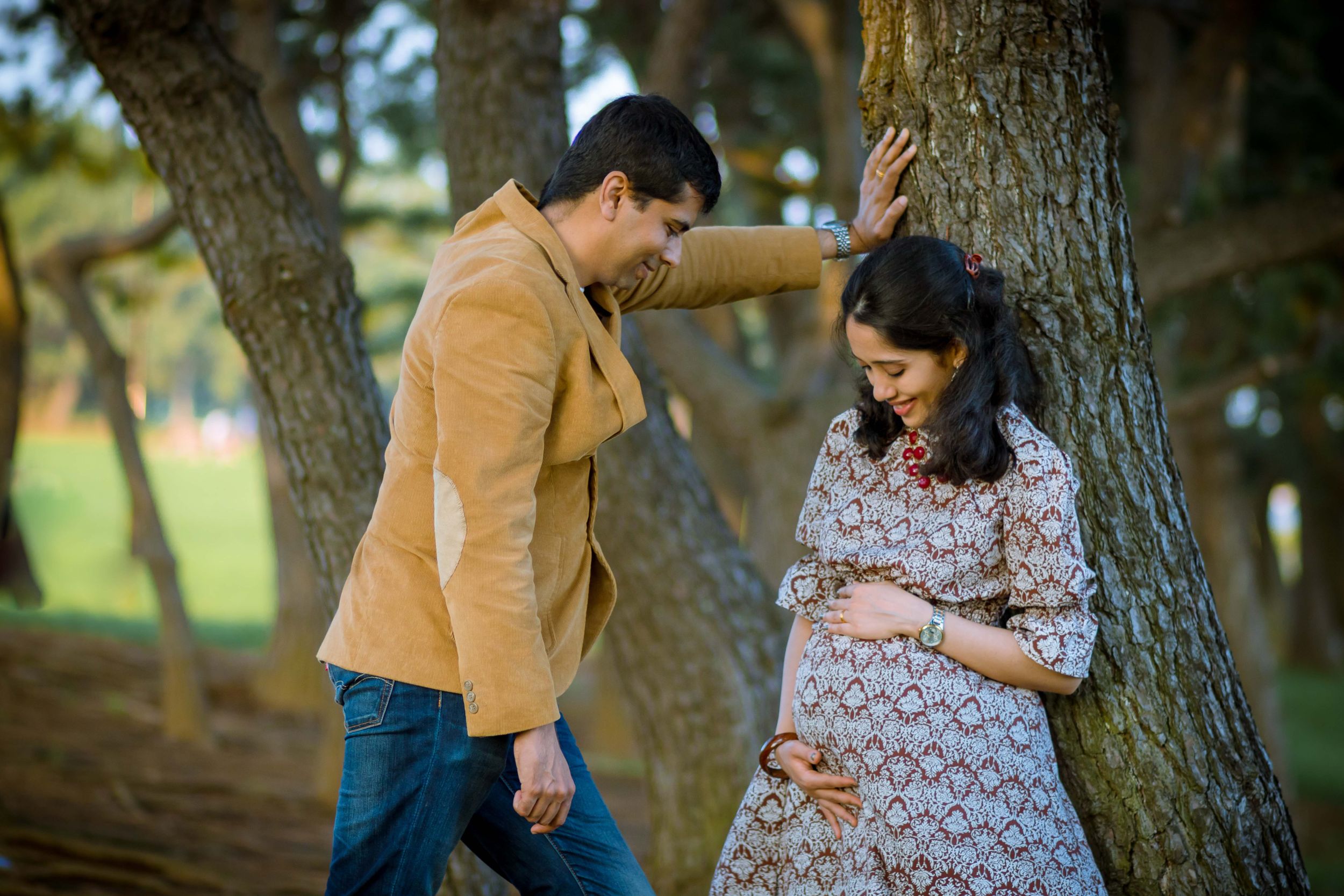 A couple shares an intimate moment in a wooded setting with the woman wearing a floral maternity dress.