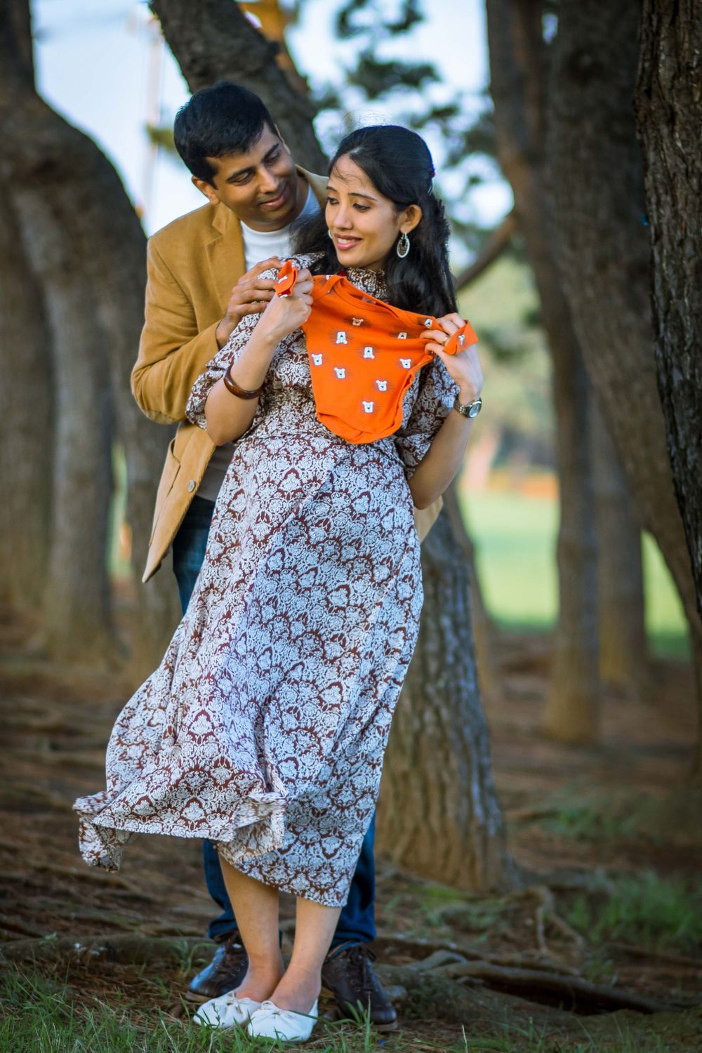 An expectant couple admires a tiny orange baby outfit while standing among trees.