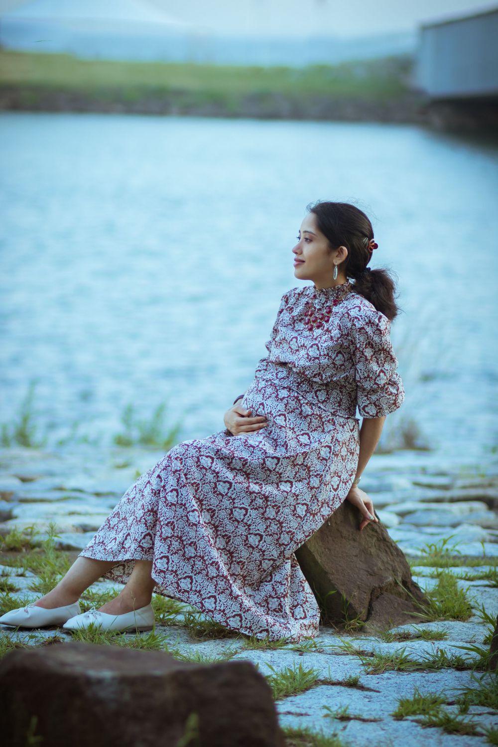 Woman in a floral dress sits peacefully by a lake on a rocky outcrop.