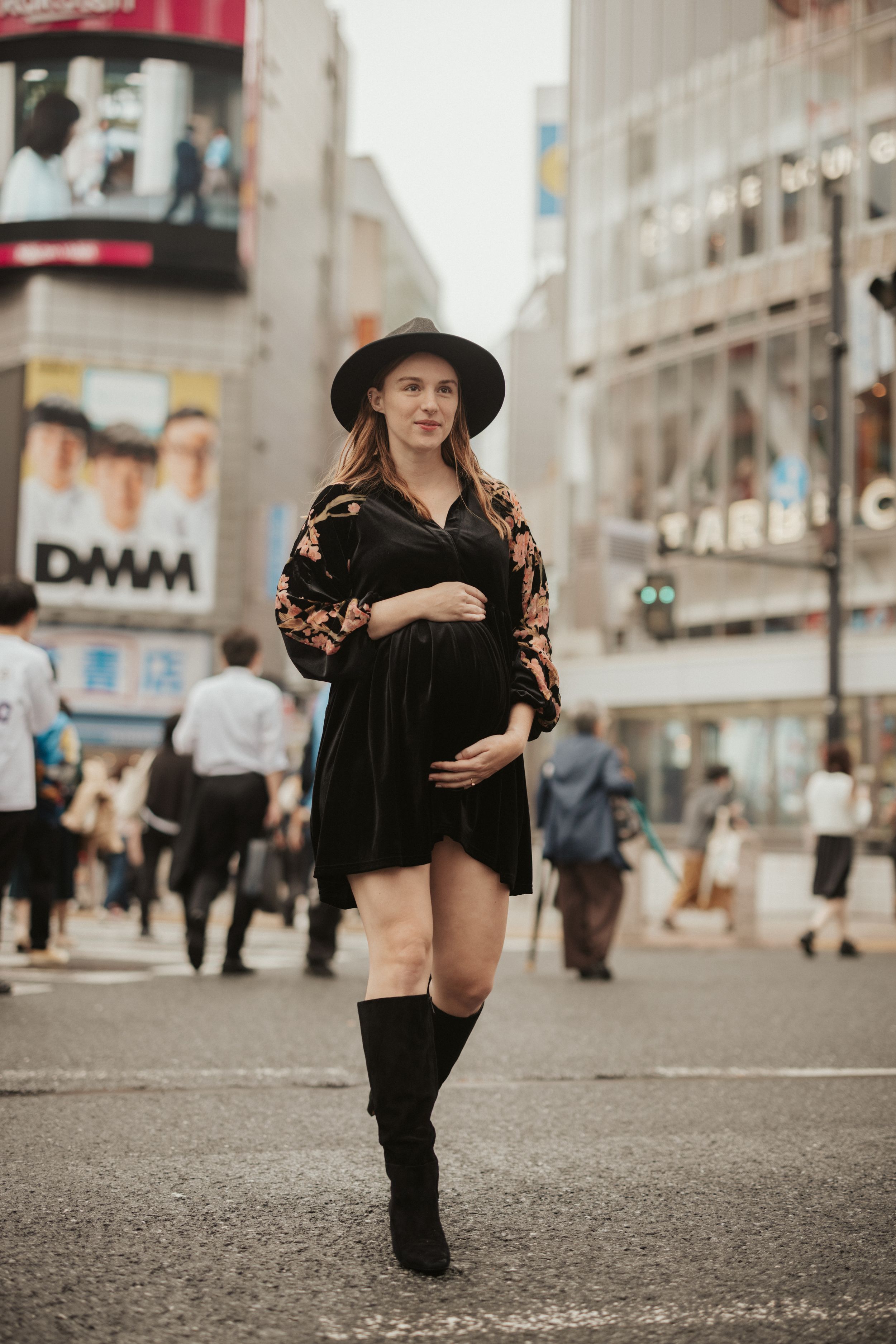 Pregnant woman in black dress and hat poses on busy city street corner with bright urban signage in background.