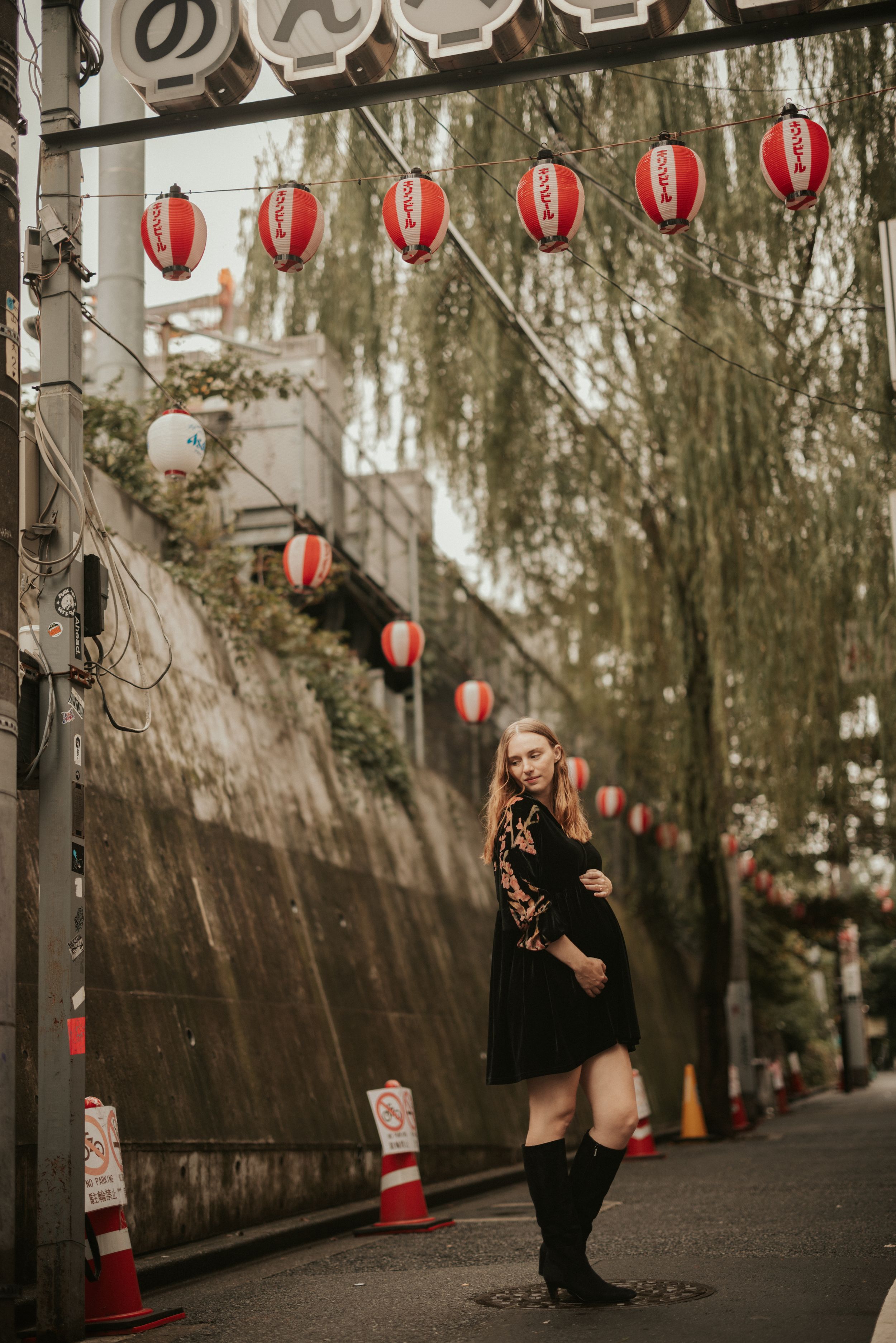 Traditional Japanese lanterns hang along a stone wall on a moody street with willow trees and evening atmosphere.