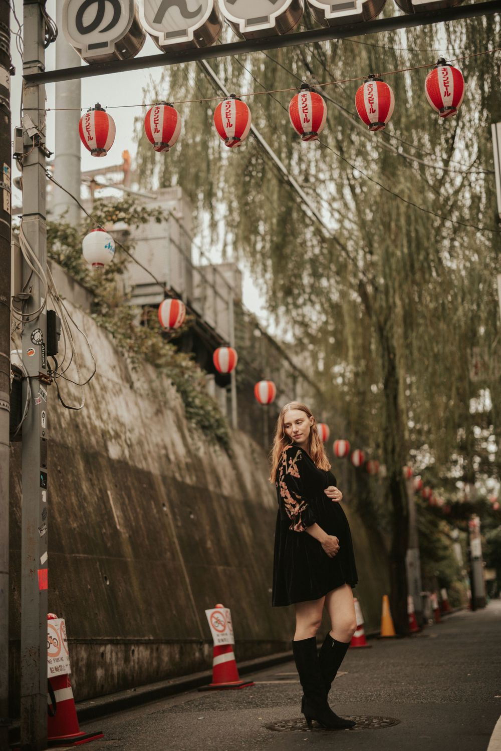 Traditional Japanese lanterns hang along a stone wall on a moody street with willow trees and evening atmosphere.