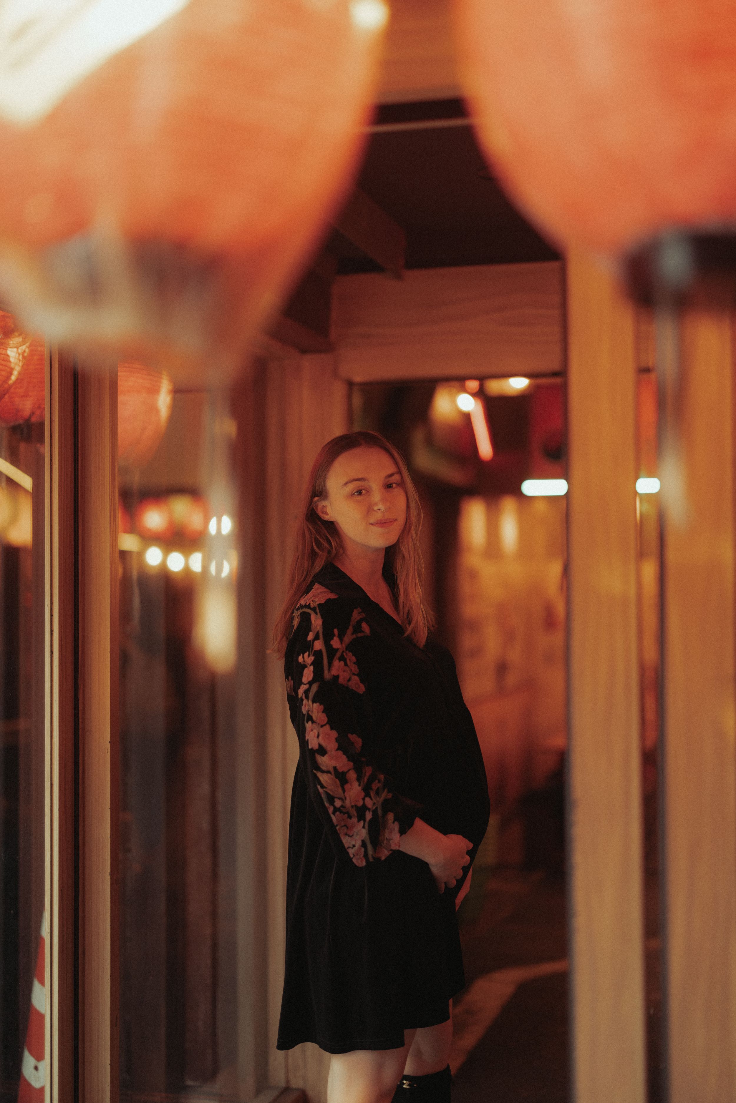 A woman in a black lace dress stands in a warm-lit corridor with glowing lights in the background.