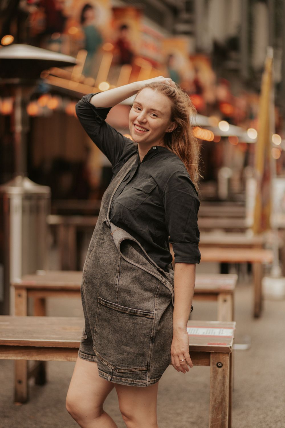 A person in a denim dress and dark shirt poses playfully at an outdoor restaurant seating area at sunset.