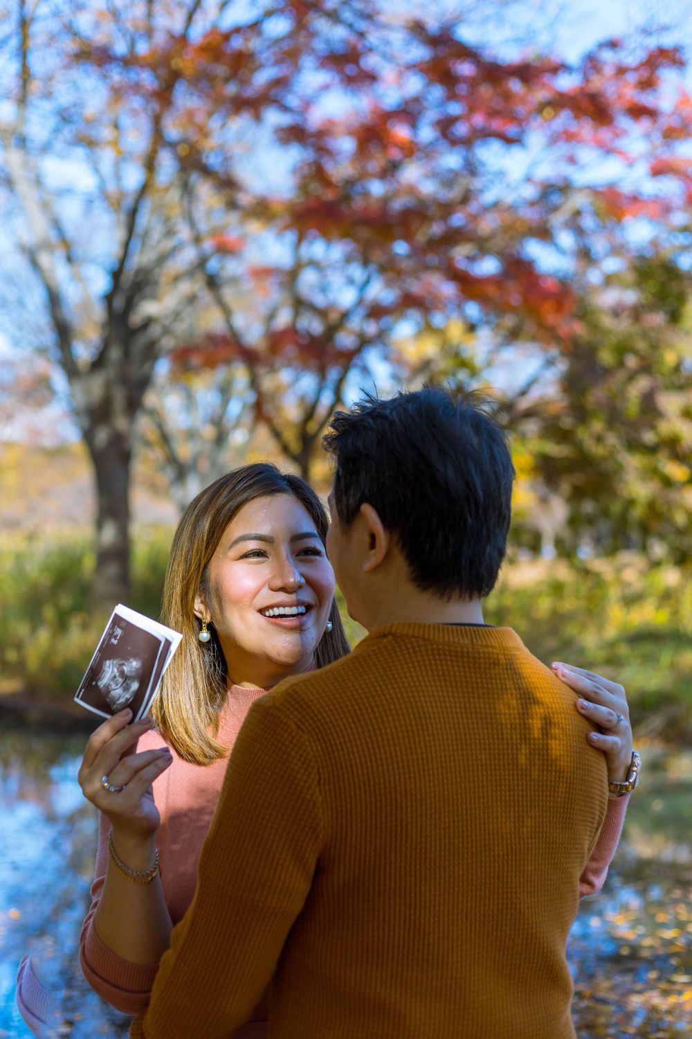 Couple sharing joyful moment among autumn trees while holding ultrasound photo.