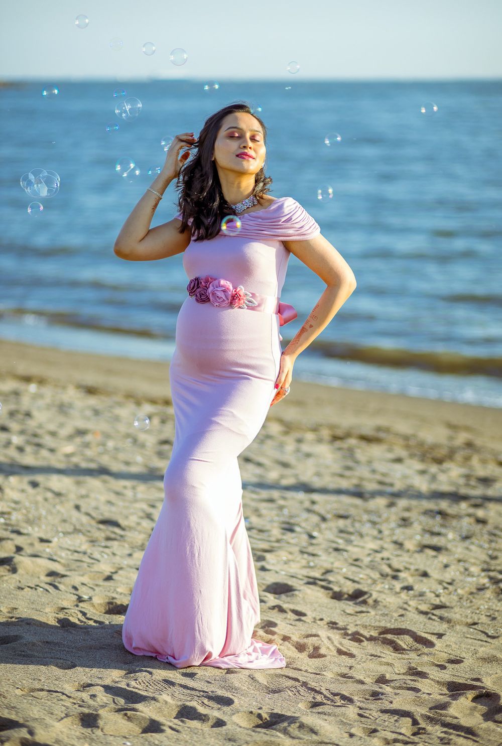 Pregnant woman in pink dress poses on beach with bubbles floating in background during golden hour.