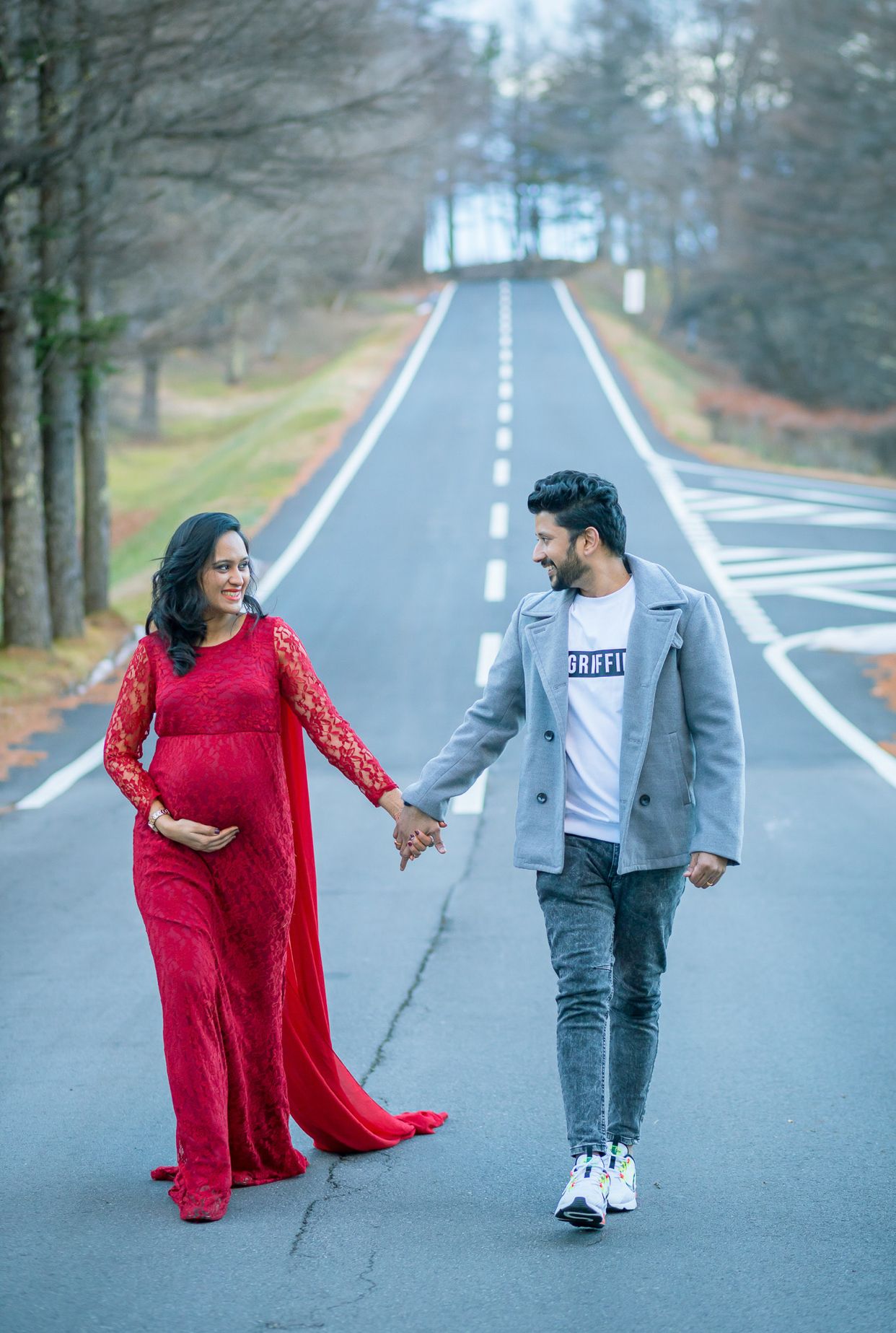 Couple walking on empty road during maternity shoot with woman wearing flowing red lace dress.