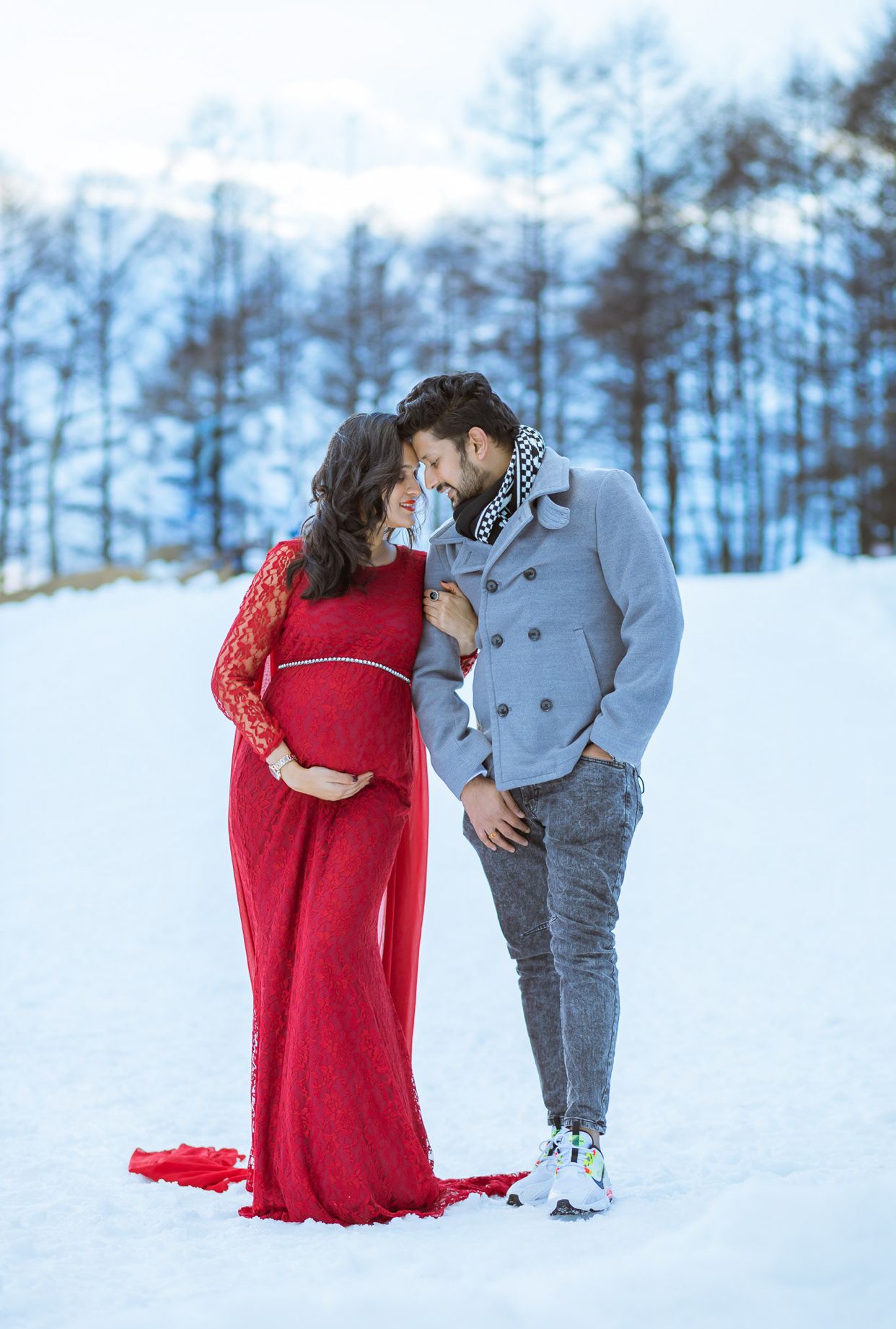 Expecting couple embracing in snowy winter landscape with pregnant woman wearing long red dress.