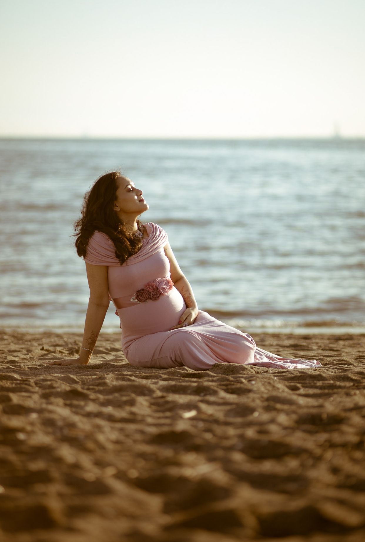Pregnant woman in pink dress relaxing on beach at sunset.