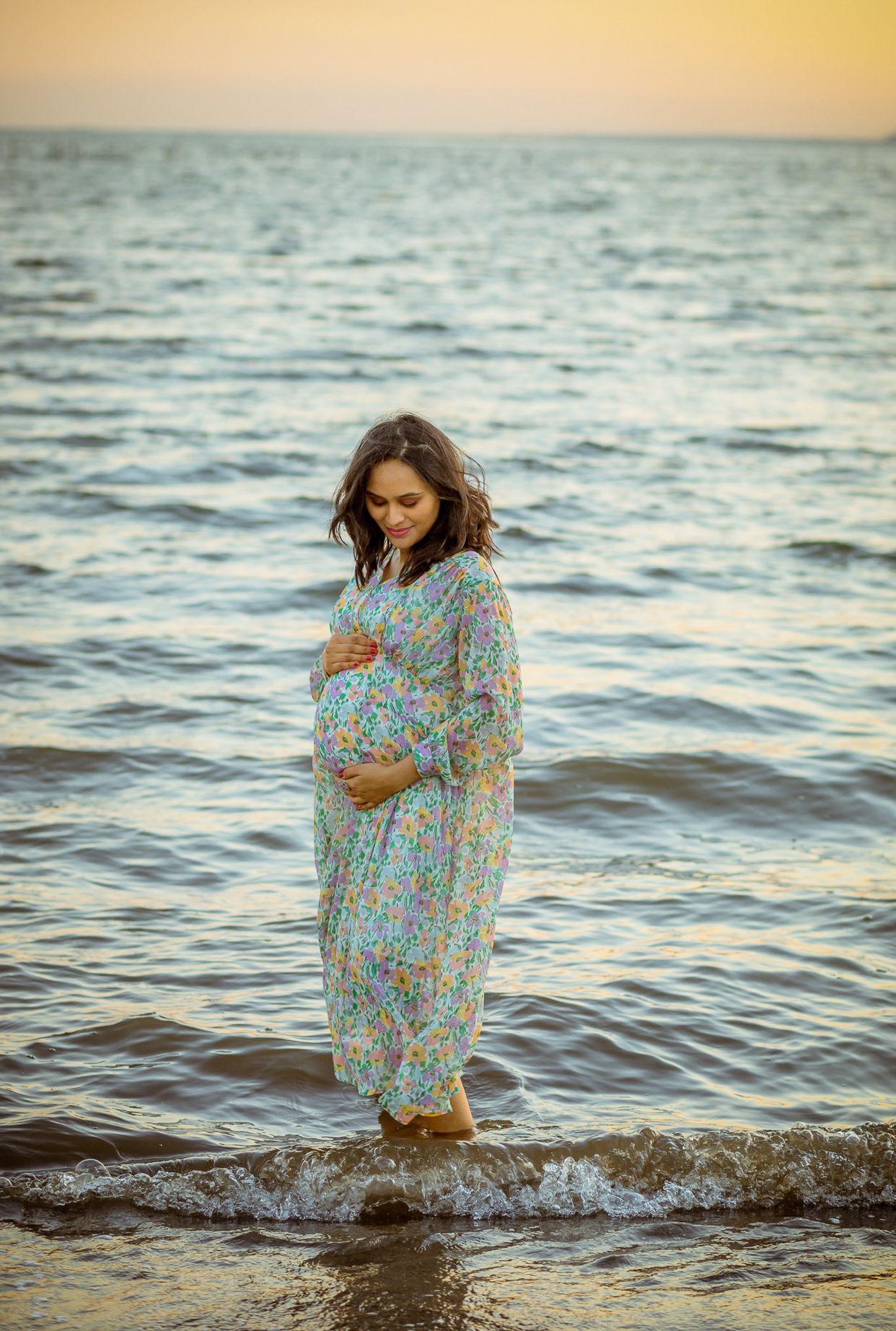 Expectant mother in floral dress standing in ocean waves during golden hour.